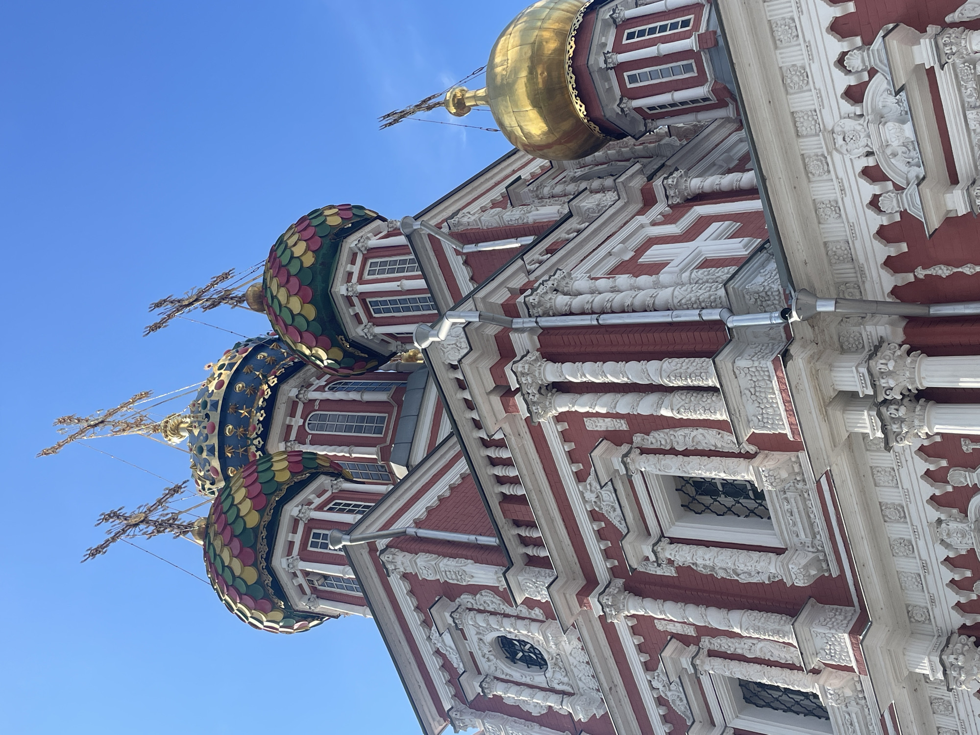 A low-angle, close-up exterior shot captures the ornate red and white facade of the Church of the Nativity of the Virgin Mary in Nizhny Novgorod, Russia. The church's architecture is characterized by intricate white stucco detailing, including columns, cornices, and decorative carvings, set against vibrant red brick walls. Several windows with decorative grates are visible. The church's domes are prominent: some are multi-colored, resembling scales in shades of red, yellow, and green, topped with elaborate gold crosses. Another dome is entirely gold, also topped with a cross. The sky above is a clear, bright blue. There are no people or animals present in the image.