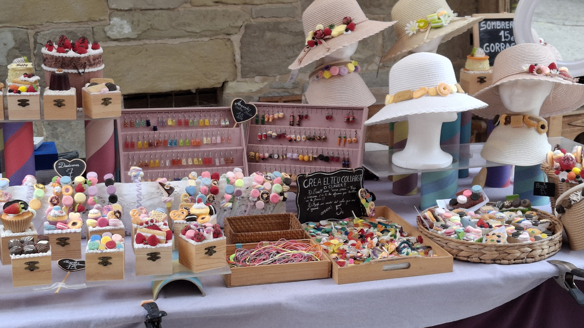 This image captures a vibrant outdoor market stall in La Fresneda, Spain, featuring a diverse array of handcrafted confectionery-themed jewelry and decorative items. The stall is arranged on a white tablecloth, with a stone wall serving as the backdrop. In the foreground, several small wooden boxes display an assortment of colorful, miniature candy-like earrings, necklaces, and bracelets, mimicking sweets such as macarons, gummy bears, and cakes. These items are meticulously arranged and appear to be made from clay or a similar material.

Dominating the mid-ground are multiple display racks filled with rows of dangling earrings. These earrings are also designed to resemble food items, including gummy bears in various colors, miniature fruits, and small pastries. To the right, several straw hats are displayed on mannequins, adorned with colorful floral and candy-like embellishments, suggesting they are also for sale. One hat features a band of colorful pom-poms, another a ribbon with cookie-like decorations, and a third, a band with what appear to be small donut charms.

A prominent sign in Catalan, "CREA EL TEU COLLARET" (Create Your Necklace), with a handwritten offer of "OFERTA! 5€" (Offer! 5€), indicates the customizable nature of some of the jewelry. Another small sign with a heart shape displays "Diademas 7" (Headbands 7). Woven baskets are also used as display containers for bulkier items like colorful threads or more assorted jewelry pieces. The overall impression is one of a whimsical and appealing craft market, with the confectionery theme creating a playful and inviting atmosphere.