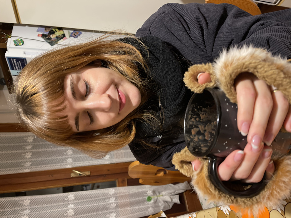 A young woman is captured in a close-up, angled shot, appearing relaxed and content indoors. She has shoulder-length, light brown or auburn hair with bangs, and her eyes are gently closed with a soft, peaceful smile on her face. She is wearing a dark, possibly black, ribbed or corduroy-like long-sleeved top.
She holds a dark-colored mug adorned with small white speckles, which is embraced by a furry, light brown cozy that resembles animal paws. Inside the mug, there is a chunky mixture, possibly oatmeal, cereal, or a thick beverage. Her hands show well-maintained nails with French tips.
The setting is an indoor domestic environment, likely a kitchen or dining area in Dosso (Ciappanico), Italy. In the background to her left, a white refrigerator is visible, decorated with various magnets, including a distinctive green owl. To her right, a wooden window frame is partially seen, covered by a sheer, white lace-patterned curtain, and part of a wooden chair. A corner of a patterned tablecloth with orange, brown, and white designs is also visible in the lower right foreground. The image evokes a sense of warmth, comfort, and quiet enjoyment. No readable text is discernible in the frame.