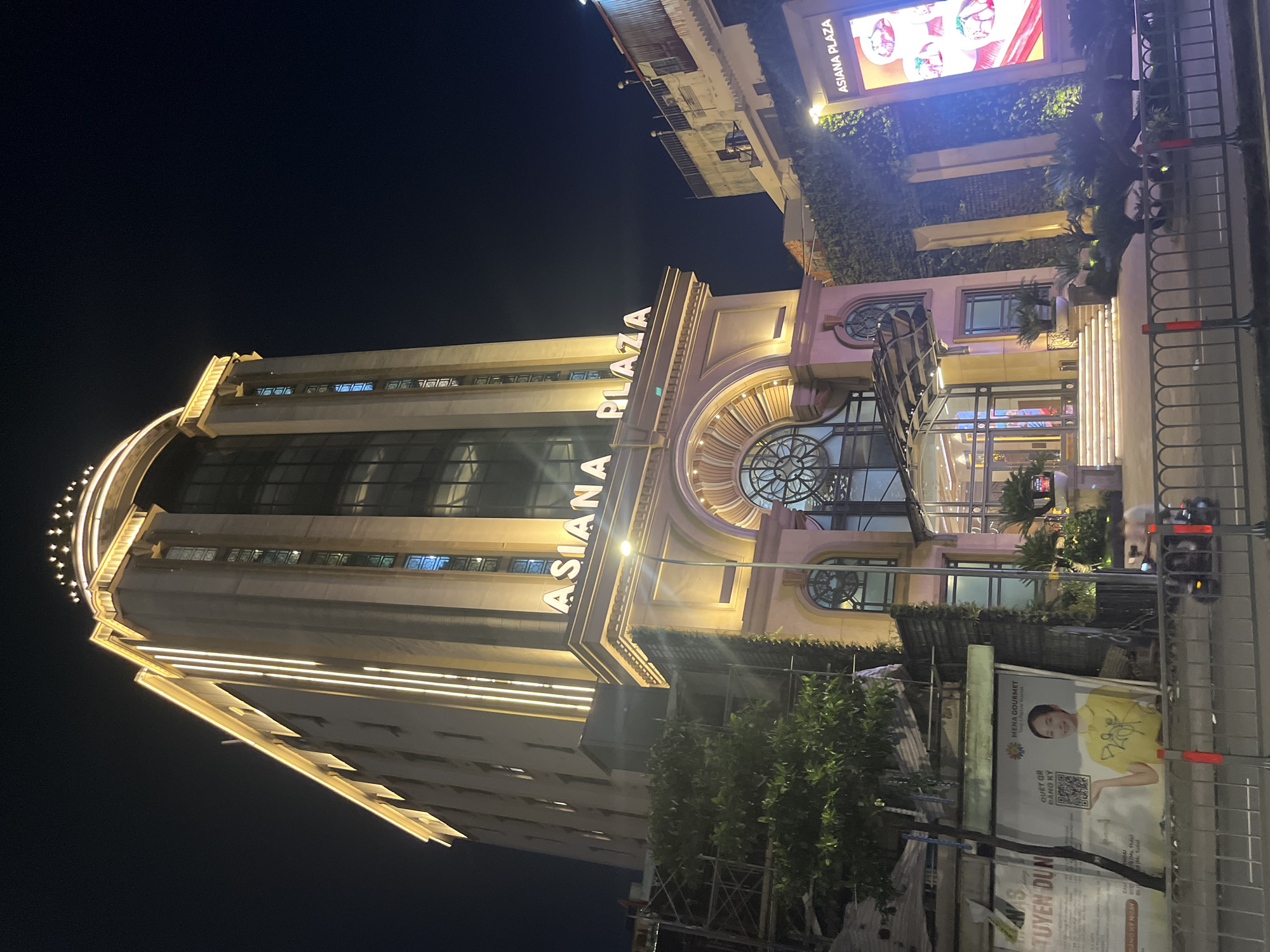 This is a vertical, eye-level, outdoor night shot of the ASIA NA PLAZA building. The building is tall and modern, with a grand entrance featuring large windows and decorative architecture. The building is illuminated by bright lights, highlighting its facade and creating a warm glow against the dark night sky. Above the building, a large billboard displays vibrant images and text. Lush green foliage adorns parts of the building's exterior, adding a touch of nature to the urban landscape. In the foreground, a black metal fence runs along the sidewalk, and a poster with a smiling person in a yellow shirt is visible. Other signs with Vietnamese text are also present. The overall impression is of a lively and well-lit commercial area at night.