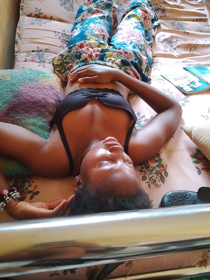 A young woman is lying on her back on a bed. She is wearing a black bra and floral-patterned pants. Her head is resting on a multicolored pillow. A book is visible on the bed to her right, along with a small black purse. The location is Umueshi, Nigeria. The bed has a floral-patterned bedspread. The view is from above, looking down at the woman.