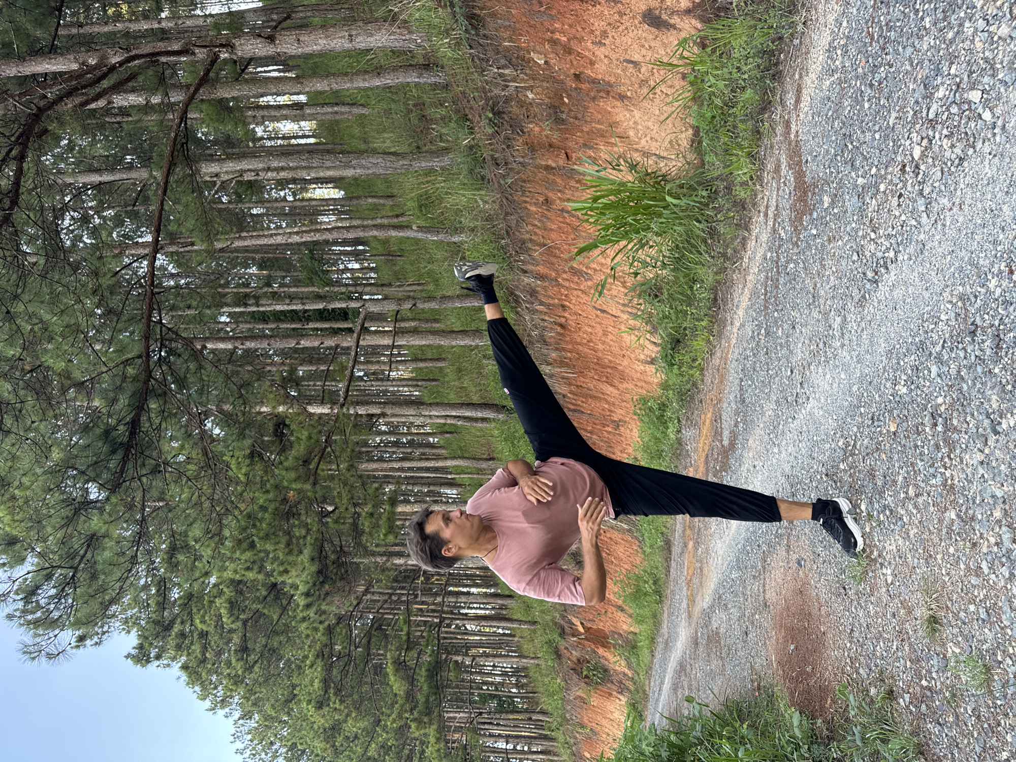 An outdoor, eye-level shot captures a person performing a standing split on a gravel path, flanked by a pine forest. The subject is a male, appearing to be in his 20s or 30s, with short dark hair. He wears a light pink short-sleeved t-shirt, black athletic pants, and black and white sneakers. His right leg is extended forward, and his left leg backward, parallel to the ground, demonstrating a high degree of flexibility. His torso is angled slightly forward and to his left, hands held near his chest, and his gaze is directed upwards.
The setting is a natural landscape, likely a forest in Da Lat, Vietnam. A grey gravel path occupies the foreground and mid-ground. To the left of the path is a reddish-brown dirt bank, partially covered with vibrant green grass and other foliage. Beyond the bank, and lining both sides of the path, are numerous tall, slender pine trees with dark green needles, forming a dense woodland. The sky, visible in the upper right corner, is clear and light blue, indicating fair weather. The lighting suggests it is daytime, possibly morning or early afternoon, with even illumination across the scene. No visible text is present.