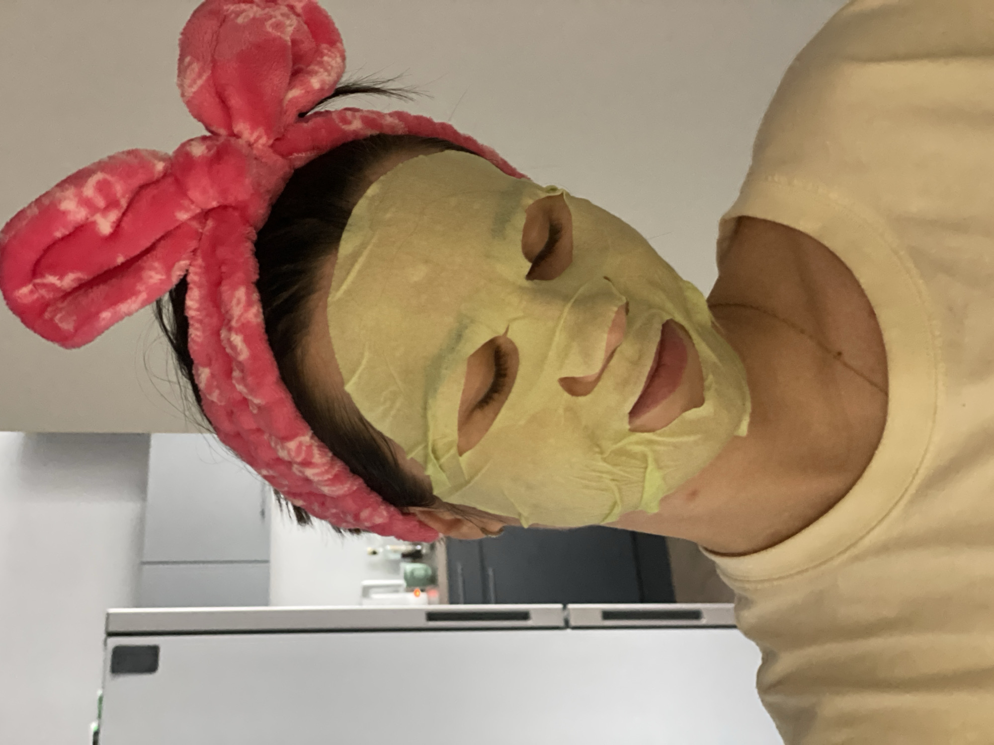 A person is wearing a sheet face mask and a pink headband with a bow. The person's eyes are closed. The person is wearing a light yellow t-shirt. The background includes a white countertop and cabinets. The location is likely indoors.
