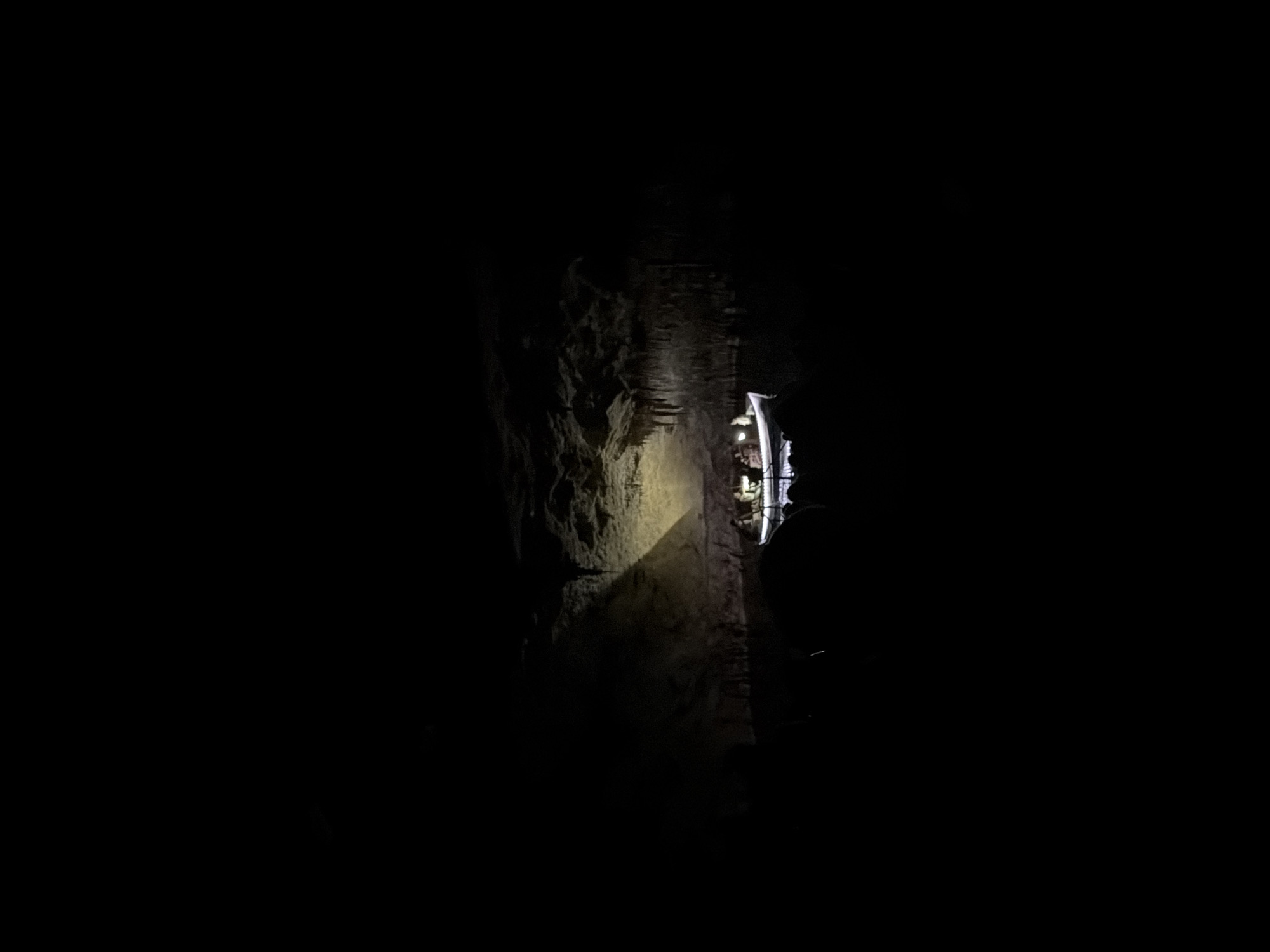 The image shows a dimly lit cave in Manacor, Spain. A guided tour boat is visible with several people onboard, illuminated by small lights. A beam of light shines on a rock wall next to the boat. Stalactites are seen on the cave ceiling. The surrounding environment is dark and rocky.