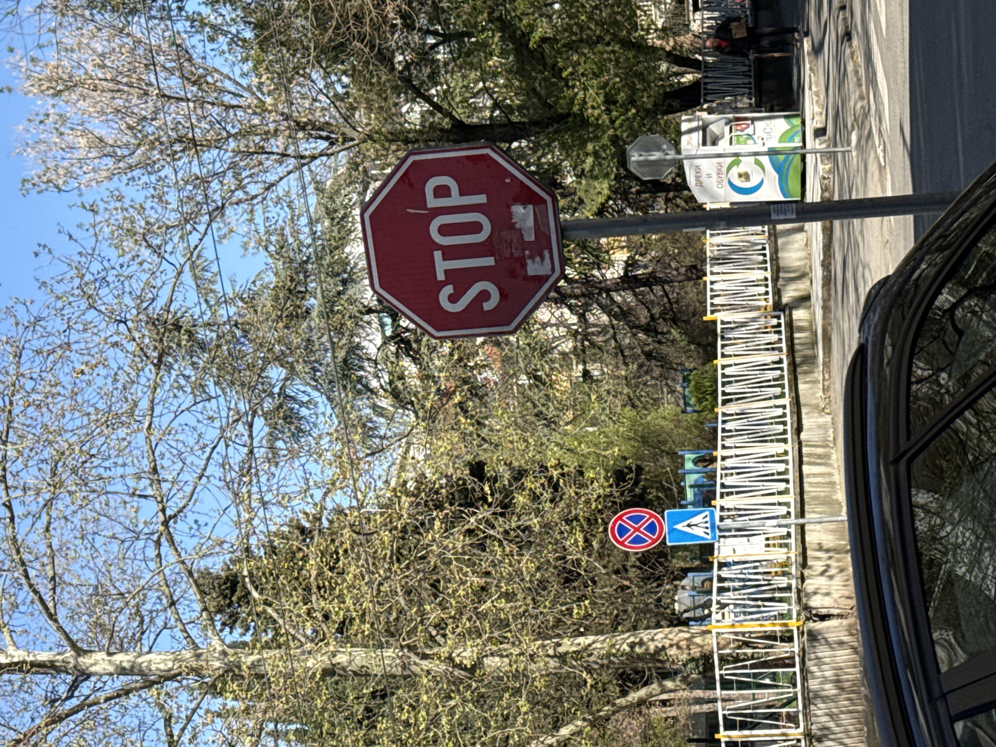 The image shows a street scene in Stara Zagora, Bulgaria. The focus is on a red octagonal stop sign with the word "STOP" in white letters. The sign is mounted on a metal pole and shows signs of wear with some peeling of the surface. Below the stop sign, partially obscured by tree branches, are two additional road signs: a round red and blue sign with a diagonal cross through it, and a blue triangular pedestrian crossing sign. A white metal fence runs along the side of the road, behind which are trees with budding leaves. A black car is partially visible on the right side of the frame. In the background, there is a recycling bin with text and images in Bulgarian. The sky is blue and visible through the trees. In the far background, people can be seen walking on the sidewalk.