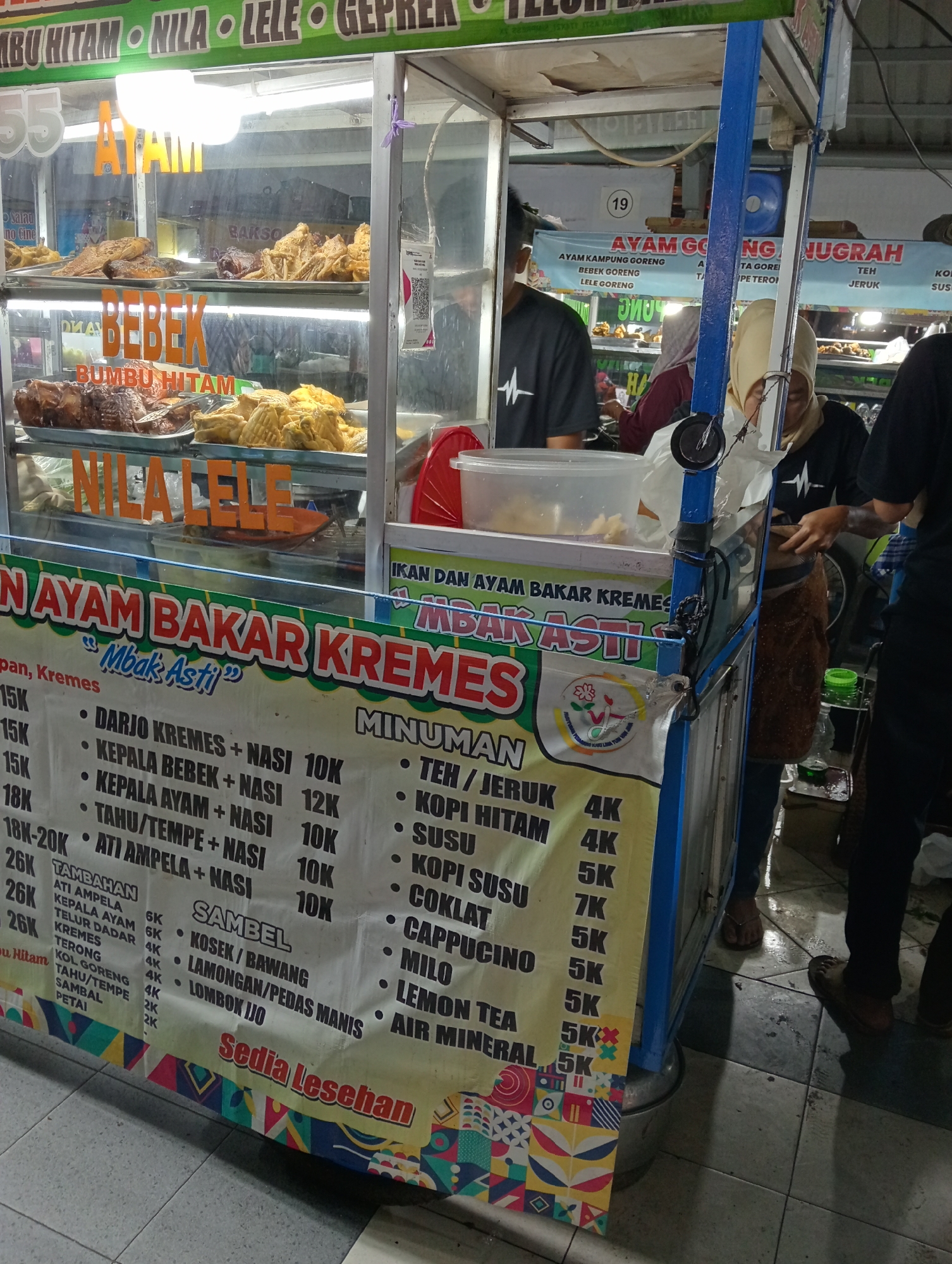 This image depicts a food stall selling various Indonesian dishes, including "Ayam Bakar Kremes" (grilled chicken with crunchy bits), "Bebek Bumbu Hitam" (black seasoned duck), and "Nila Lele Geprek" (spicy fried tilapia). The stall is brightly lit and appears to be in an outdoor setting, possibly a street food market. A banner displays a menu with prices for different meal combinations and individual items. Several people are visible, including vendors and customers, some of whom are preparing or serving food. The stall is decorated with colorful banners and signage, typical of food businesses in Indonesia. The overall impression is that of a busy and vibrant food establishment.