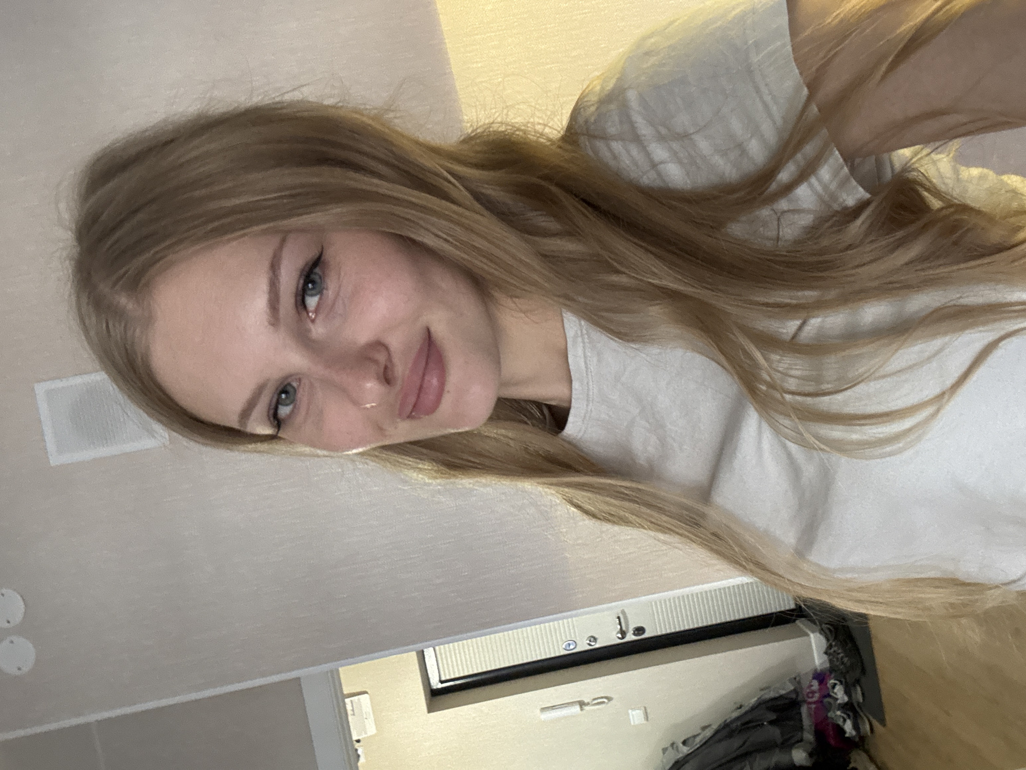 The image is a close-up selfie of a young Caucasian woman with blonde hair and blue eyes. She is wearing a light grey t-shirt and a small nose ring. She is indoors, in what appears to be an apartment hallway. To the left of the woman, a door is visible, with a light-colored wall and a wooden floor. The wall has a light texture. There are some items of clothing and shoes on the floor near the door. The lighting in the image is warm and slightly dim. The overall impression is a casual, personal snapshot.
