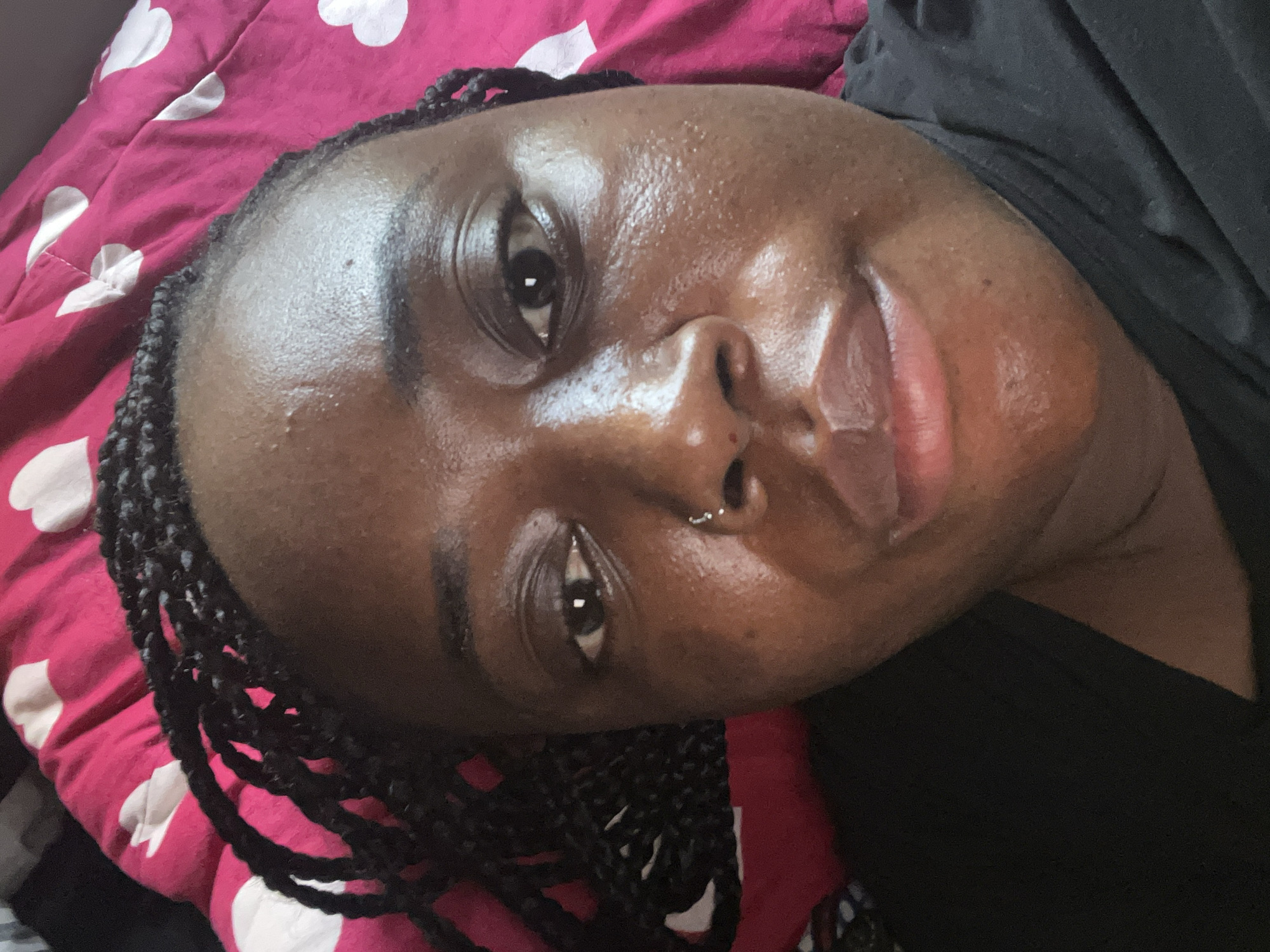 A close-up, angled portrait shows a person, likely a woman, with dark skin and dark braided hair. Her head is positioned on or against a pink bedsheet or blanket adorned with a pattern of white hearts. She gazes directly at the camera with a calm, gentle expression and a slight, soft smile. Her skin has a noticeable sheen, particularly on her forehead, cheeks, and nose, suggesting it is moisturized or oily. A metallic septum ring is visible piercing her nose. She wears a dark-colored garment, possibly a black top or t-shirt, visible on her shoulder and upper chest. The lighting is soft and even, characteristic of an indoor setting. No specific activity beyond posing for the camera is discernible, implying a relaxed, casual moment. The location context for this image is Abuja, Nigeria.