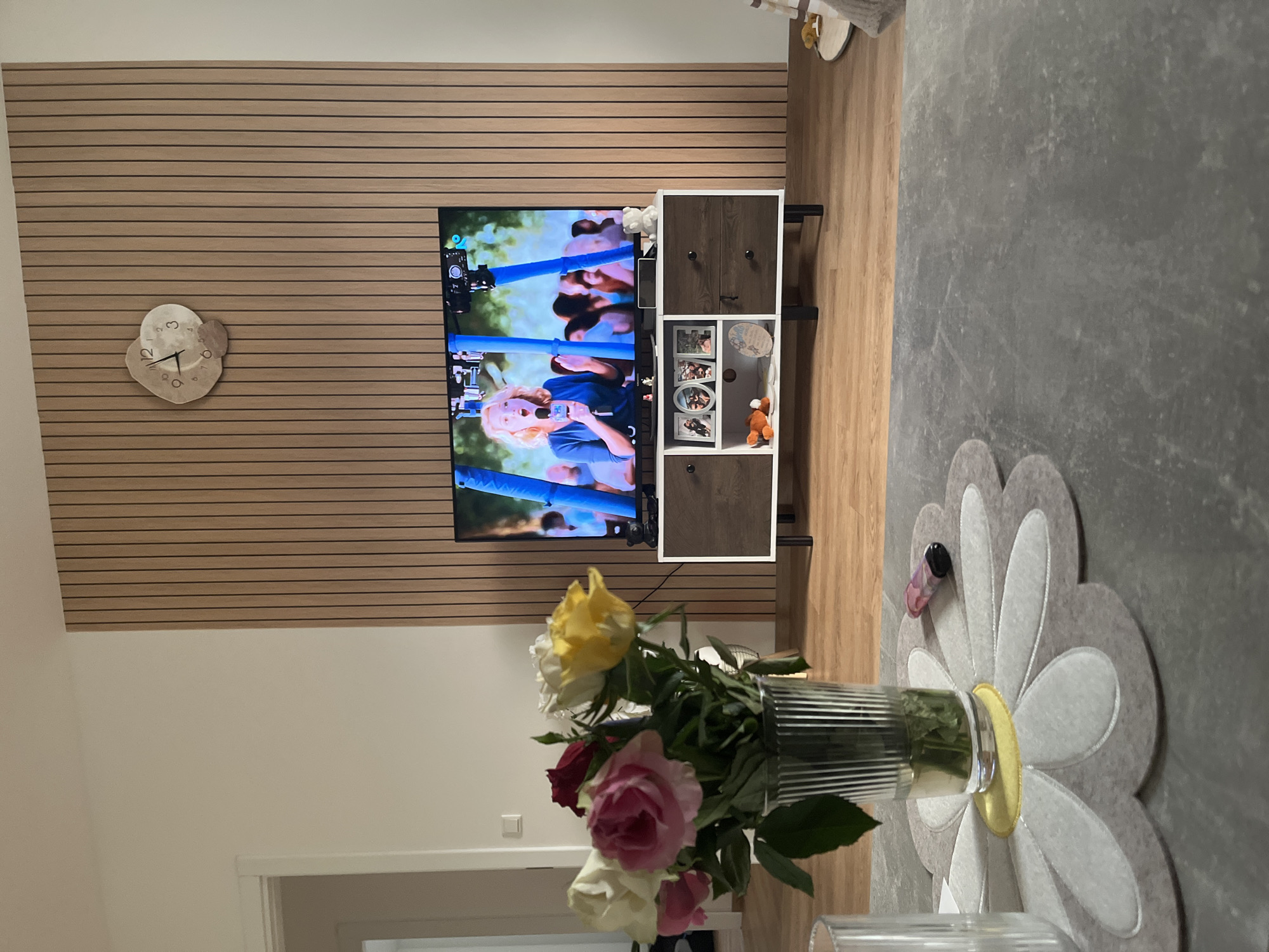 The image depicts a living room scene, focusing on a television that is on and displaying a woman speaking into a microphone. The television is placed on a media console. Above the television, on a wall adorned with vertical wooden panels, hangs a decorative clock. In the foreground, on a grey table, is a vase filled with a bouquet of roses of various colors. The room appears to be well-lit, with a light-colored wall visible on the left. The overall impression is that of a modern, cozy living space. The image does not contain any information that would allow for an identification of the location as Garmisch-Partenkirchen, Germany.