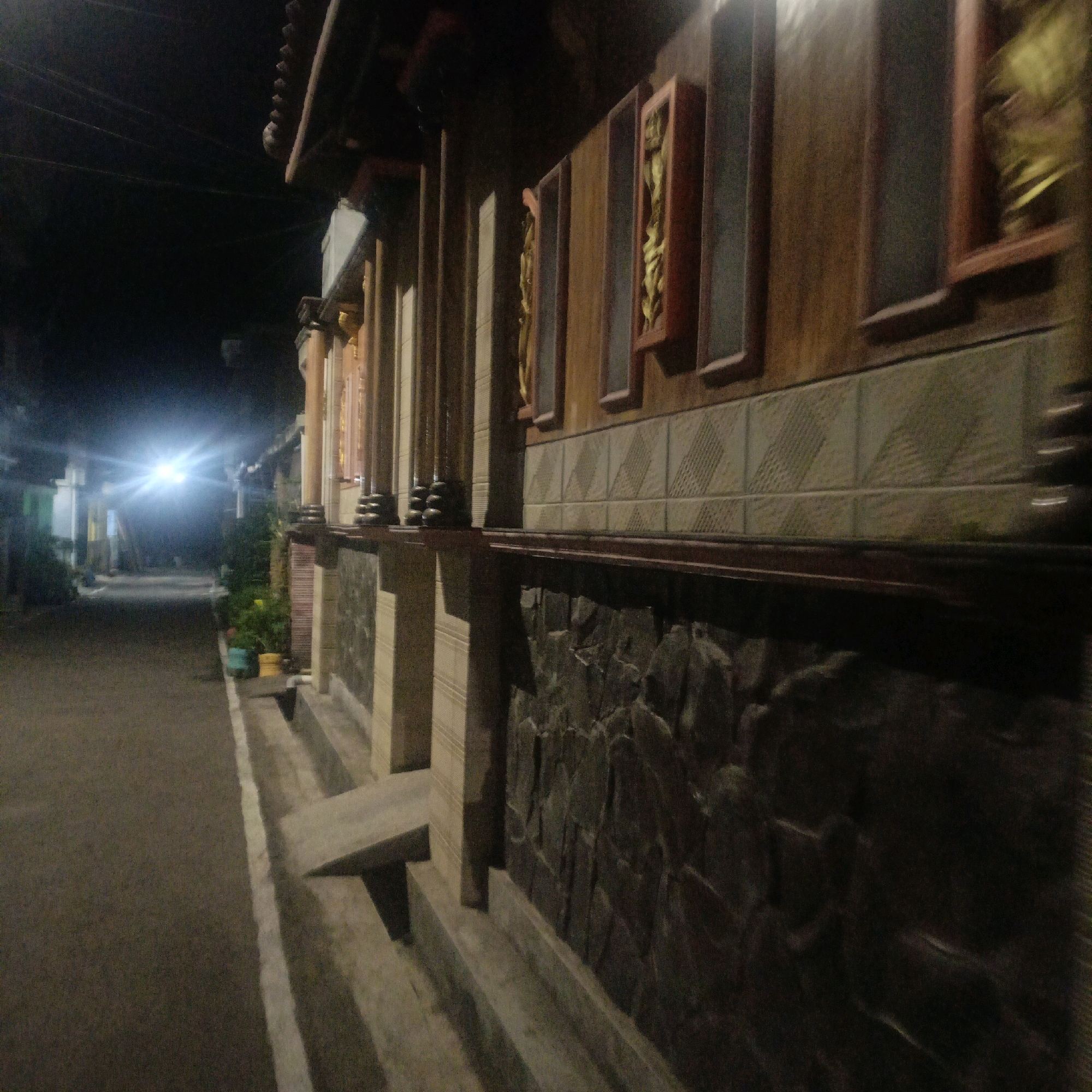 The image depicts a nighttime scene on a narrow, paved street in Salatiga, Indonesia. The street is lined with a building featuring ornate architectural details. The building's facade is adorned with decorative tiles, stone accents, and recessed wooden frames containing gold-colored decorative elements. The foreground shows a concrete walkway with steps leading up to the building. The street is illuminated by a bright, almost blinding light emanating from the distance, suggesting a powerful light source like a streetlamp or vehicle headlights. The surrounding environment is dark, with faint outlines of other buildings and vegetation visible in the background. There are no people or animals clearly visible in the image.