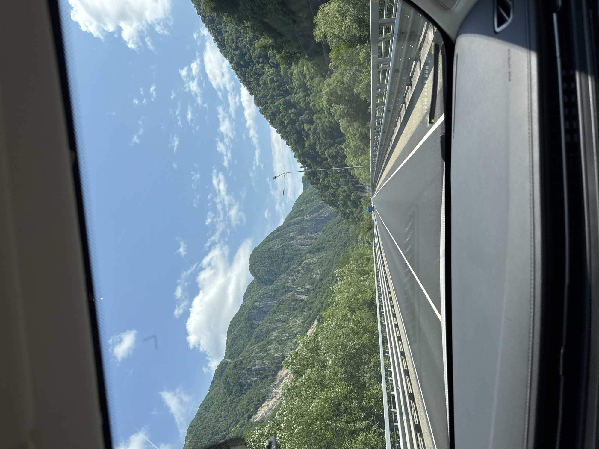 The image shows a view from inside a car driving on a highway through a mountainous, forested region. The highway is paved with asphalt and has white lane markings. It is bordered by a metal guardrail on both sides. Lush green trees cover the steep slopes of the mountains that flank the highway. The sky above is blue with scattered white clouds. A few streetlights are visible along the side of the road. The dashboard of the car is visible in the foreground. There are no people visible in the image, and no specific landmarks are identifiable that would suggest the location within the city of Monastyr, Russia, as the image depicts a natural landscape outside of a city.