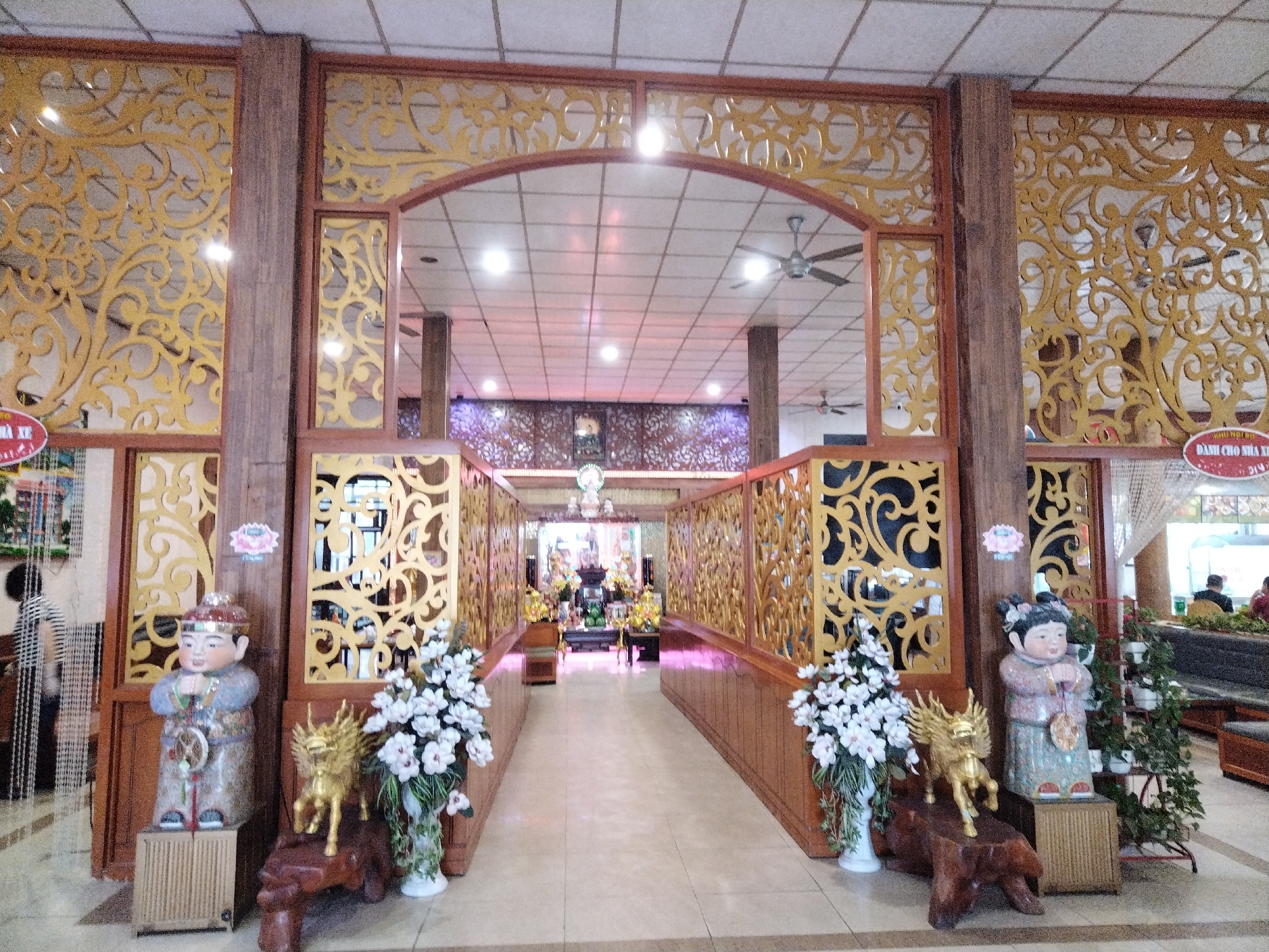 The image depicts a view of the interior of a building, possibly a temple or religious structure, located in Chu Văn An Ward, Vietnam. The scene is framed by an elaborate wooden entryway with ornate gold-colored filigree designs. Two child-like statues stand on either side of the opening, along with flower arrangements, a sculpture of a golden kirin and various potted plants. The statue on the left is a male figure and the statue on the right is a female figure.
The interior beyond the entryway features a long corridor with similar wooden dividers and floral decorations. In the distance, religious icons and objects are visible. The ceiling is lined with fluorescent lights. The building appears to be well-maintained and illuminated, suggesting active use and care. The setting includes tiled floors, and the space is open and seemingly spacious.
A few people are visible in the far background, near what appears to be the entry to the building behind the ornate decorations.
