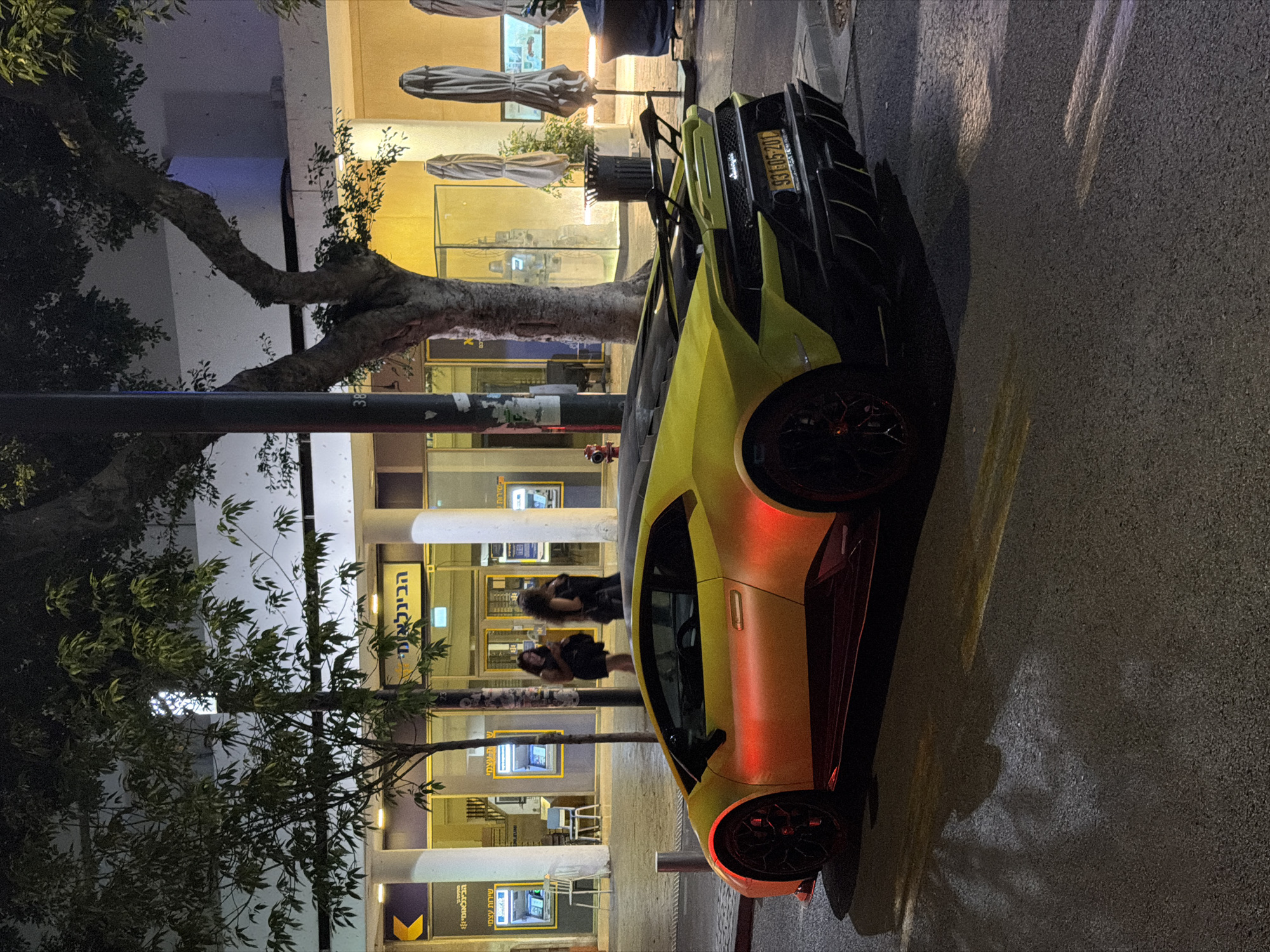This nighttime image depicts an urban street scene in Tel-Aviv, Israel. A vibrant yellow Lamborghini sports car, likely a Huracan model, is prominently parked on the asphalt street, angled slightly towards the left. The driver's side of the vehicle, particularly the front fender and door, exhibits a striking orange-red illumination, contrasting with the car's primary yellow paint. The car features dark red/maroon side skirts and black multi-spoke wheels with visible red brake calipers. An Israeli license plate, "93-115-20", is affixed to the rear, with the city name "TEL-AVIV" legible beneath it.
In the background, a commercial building complex is illuminated. On the left, the facade of a branch of "The First International Bank of Israel" (הבינלאומי) is visible, featuring pillars and multiple self-service ATMs behind glass, marked with "שירות עצמי" (Self Service). Two women are present near the ATMs; one, in dark attire, appears to be using a mobile device, while the other stands slightly behind her. To the right, obscured by a prominent tree, is an area that appears to be an outdoor seating establishment, marked by several closed, folded beige umbrellas and a black public trash can. Further right, a large glass display case, possibly containing an art installation, reflects light. A red fire hydrant is partially visible on the far left, near the street curb. Various trees with dark foliage frame the scene, and artificial lighting from the buildings casts warm yellow and orange hues across the environment and onto the wet or reflective street surface.