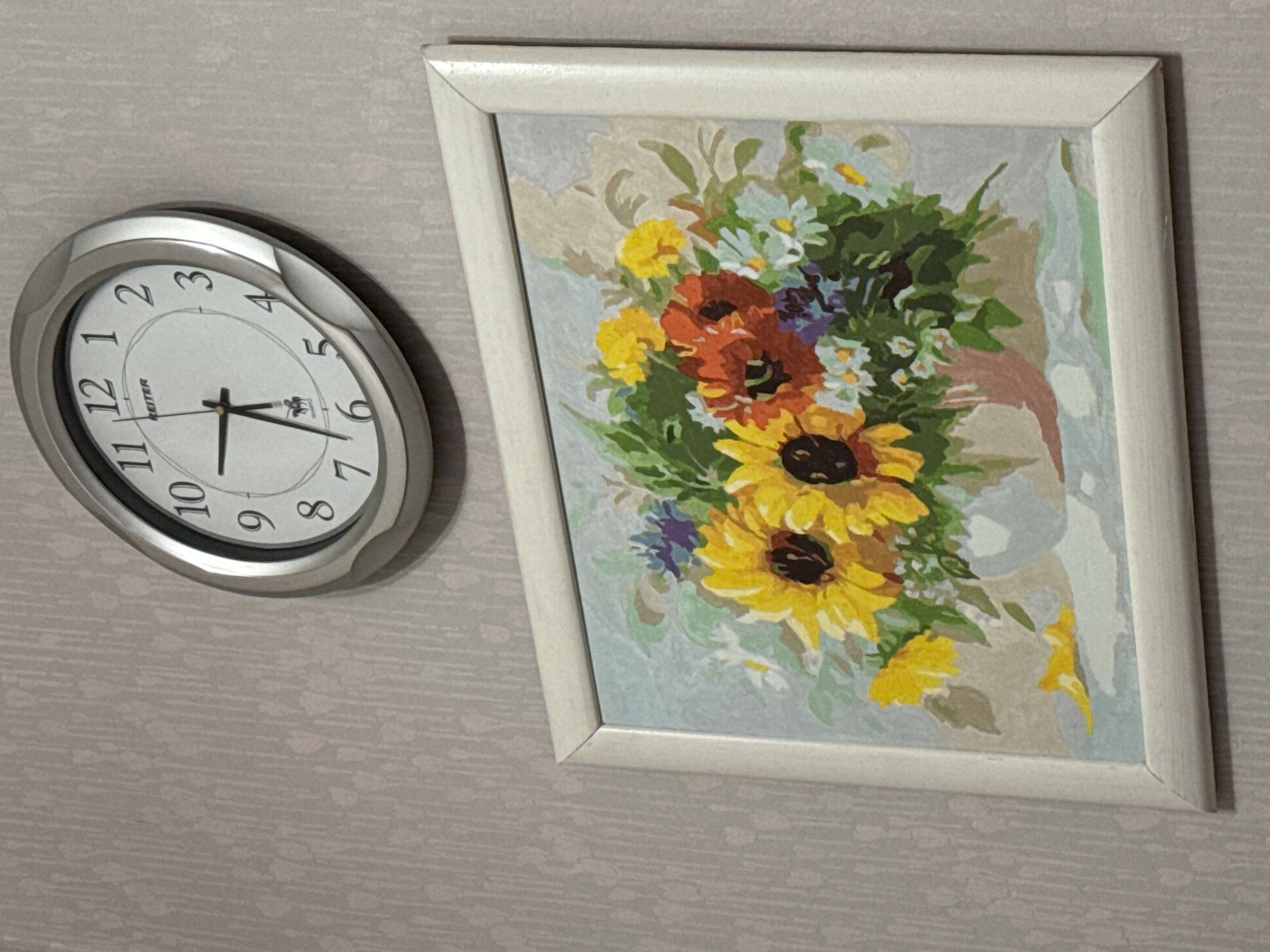 The image shows a wall clock and a framed painting of flowers. The wall clock is circular with a silver frame and a white face with black numbers and hands. The painting is rectangular with a white frame and depicts a bouquet of sunflowers and other flowers in a vase. The background of the image is a textured wallpaper. There are no people or animals visible in the image. The location of this scene in Nizhny Novgorod, Russia, cannot be determined from the provided image.