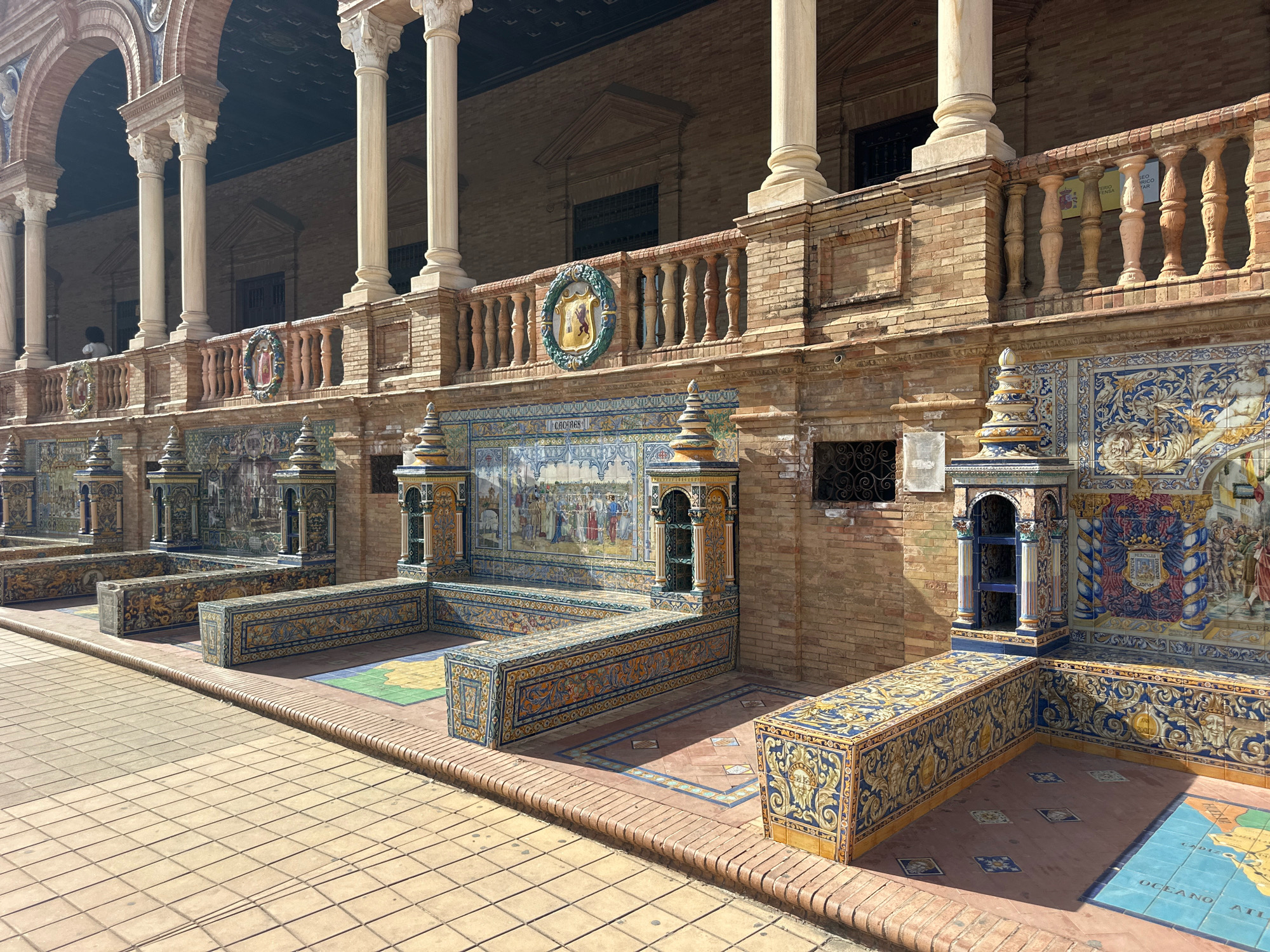 The image shows an outdoor courtyard area with ornate tiled benches and a brick building with arched walkways supported by columns. The tiles feature intricate patterns in blue, yellow, and white, as well as pictorial scenes. The ground is paved with square tiles. In the background, a person is visible standing on a balcony area. The overall architecture and tilework suggest this is likely the Plaza de España in Seville, Spain.