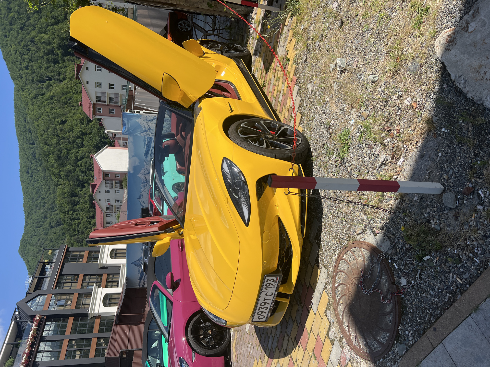 In the city of Estosadok, Russia, on a sunny day, a vibrant yellow convertible sports car with its dihedral doors open is parked on a colorful patterned pavement, bordered by gravel. The car displays a Russian license plate, "0939TC 193". Behind the car, several other luxury convertibles in pink and turquoise are visible, along with a backdrop of modern multi-story buildings and lush green mountains. A red chain secured by stanchions cordons off the area in front of the car. In the foreground, a manhole cover with a chain draped over it lies on the gravel.
