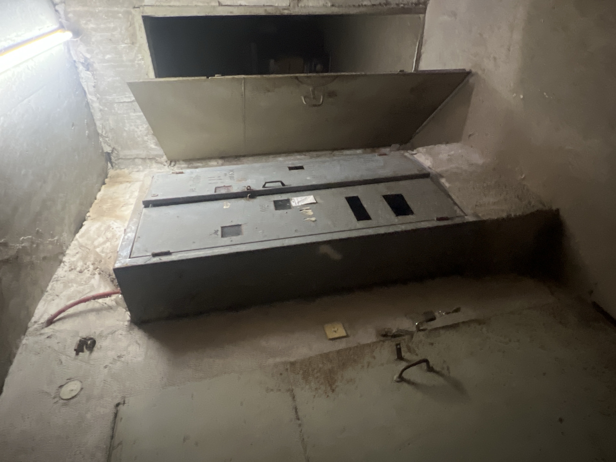 The image depicts a utility access point, possibly within a building in Elena, Bulgaria. A partially open, hinged metal door reveals an interior space containing a metal electrical box. Various pipes and electrical conduits are visible on the surrounding wall. The overall impression is one of aged, functional infrastructure. No people are present.