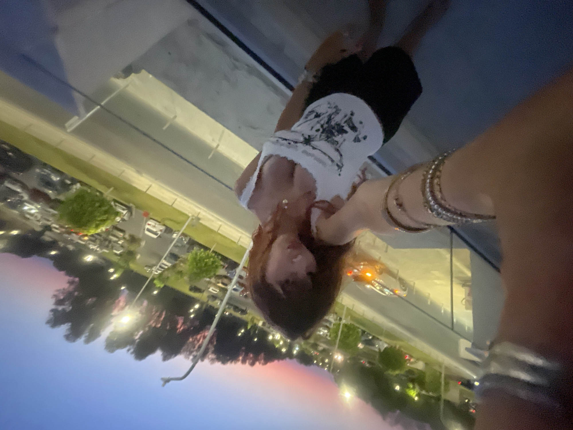 The media file shows a person, likely a woman with long, reddish-brown hair, taking a selfie at dusk. She is wearing a white tank top with a black graphic print and black shorts. She has several bracelets on her arm. The setting appears to be an elevated outdoor area, possibly a balcony or rooftop, overlooking a parking lot filled with cars. Streetlights and the headlights of cars are visible, indicating it's nighttime. The sky displays the soft colors of twilight, with hues of pink, orange, and blue. The overall mood seems casual and relaxed, perhaps during a social outing or travel. No visible text is present. The location context is Paphos Municipality, Cyprus.