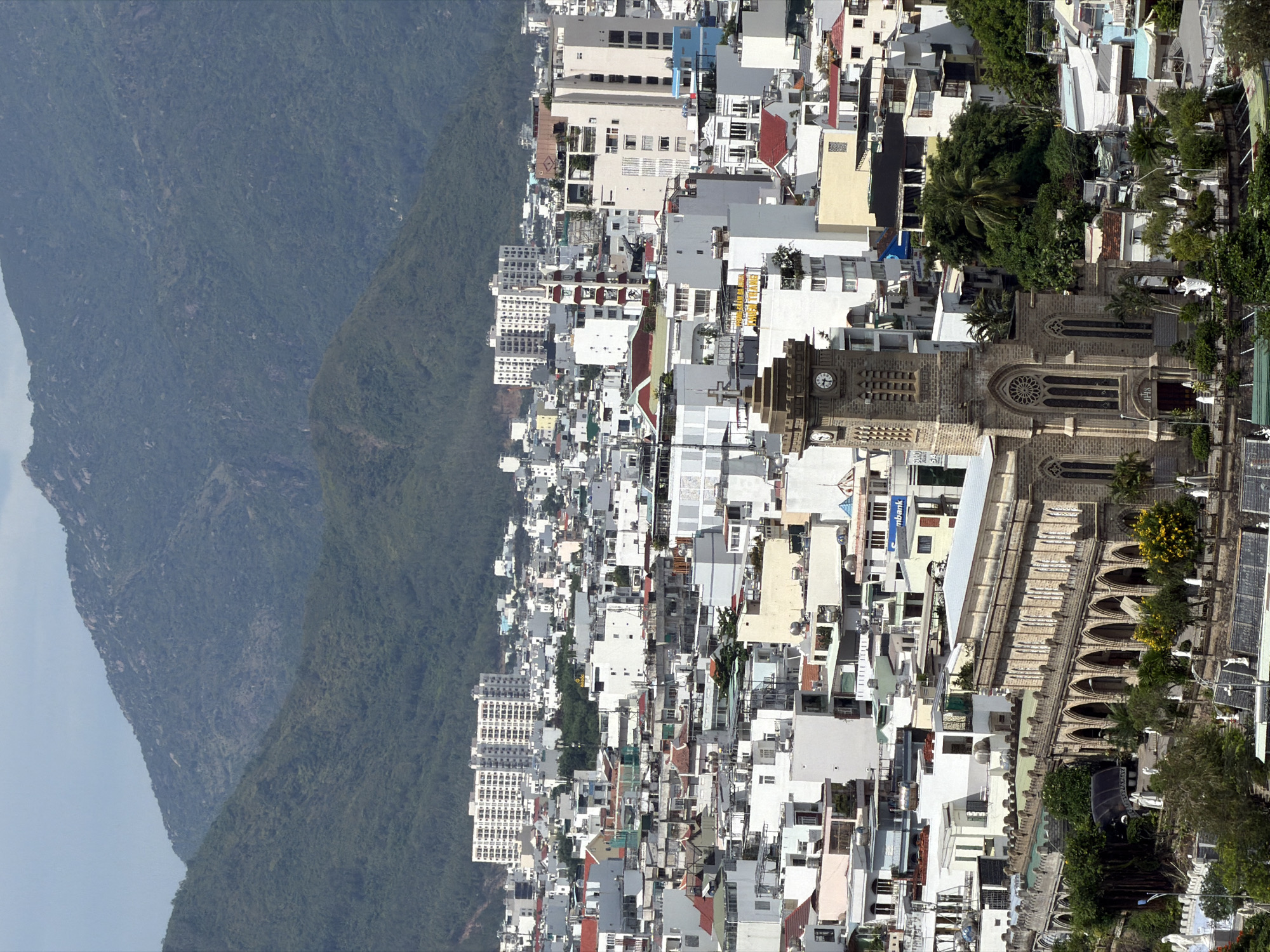 A panoramic photograph captures a vibrant cityscape nestled against a backdrop of majestic, lush green mountains under a bright, clear sky. The city, which fills the foreground and mid-ground, is densely packed with numerous buildings of varying heights and architectural styles. Many structures are white or light-colored, featuring a mix of flat and sloped roofs in shades of red, grey, and brown.
A prominent feature in the right-center of the image is a large, grey stone church, characterized by its Gothic architecture. It boasts a tall clock tower with a visible clock face displaying approximately 10:20. The church also features pointed arched windows, a distinctive rose window, and architectural buttresses along its length. The letters "IHS" are inscribed above its main entrance.
Scattered throughout the urban landscape are numerous trees, including several palm trees, providing pockets of natural greenery amidst the concrete. In the background, dark green, densely forested mountains rise dramatically, their rugged contours forming a stark contrast to the sprawling urban development. The clear, bright sky suggests a sunny late morning or early afternoon.
No discernible people or specific activities are visible in the scene. Visible text includes "PHUONG TRANG" on a yellow building within the city and "VPBank" on another building located to the left of the church. Solar panels are also visible on the roofs of some buildings.