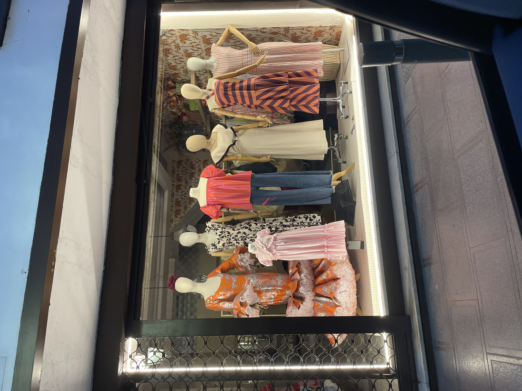 The image shows a storefront window displaying several mannequins dressed in various outfits. The storefront is located on a street with paved sidewalks and a streetlight visible. The window features a display of clothing on mannequins, including a ruffled orange dress, a pink gingham dress, a black and white floral dress, a striped orange and purple dress, and a pink striped dress. There are also mannequins wearing blouses and pants. The background of the display appears to be wallpaper with an elephant pattern. The lighting in the scene is a mix of interior store lighting and ambient street light. The overall impression is that of a clothing boutique display.