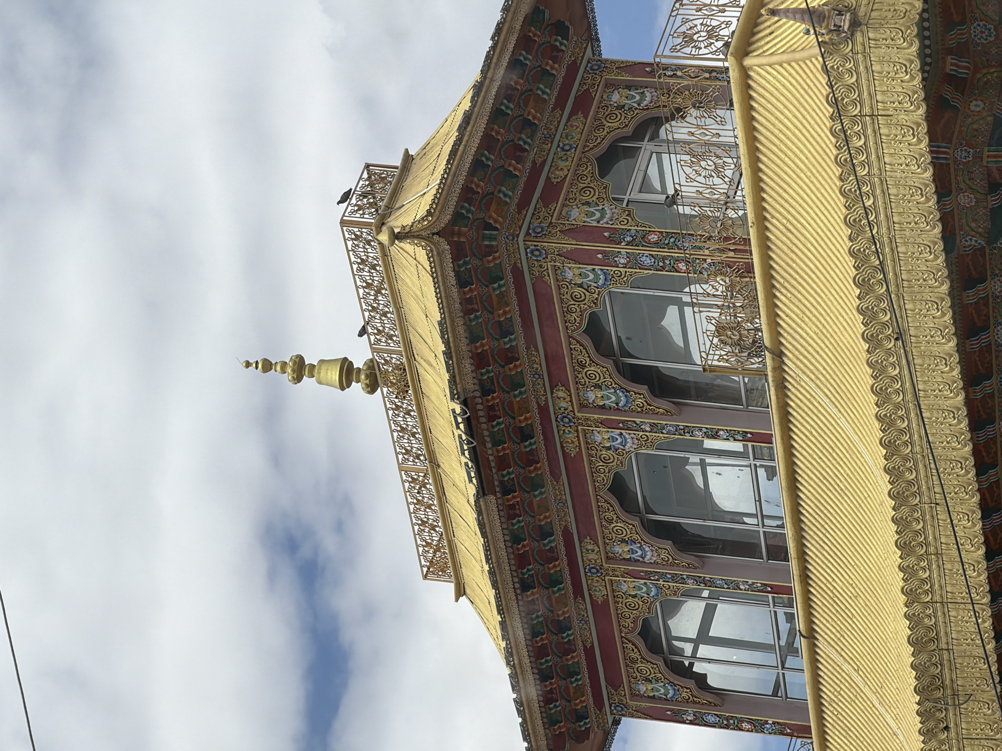 The image shows the roof and upper levels of a building in Leh, India, likely a monastery or temple, with a golden roof and ornate decorations. The architectural style is distinct, featuring elaborate carvings and paintings in vibrant colors, primarily red, gold, blue, and green. The building has large arched windows with reflective glass. A golden spire tops the roof. Several pigeons are visible perched on the roof and a decorative metal railing. The sky is partly cloudy with scattered white clouds against a blue background. A thin, dark wire runs across the upper left portion of the image, possibly a power line or antenna. The overall scene depicts a well-maintained and culturally significant structure within the city of Leh.