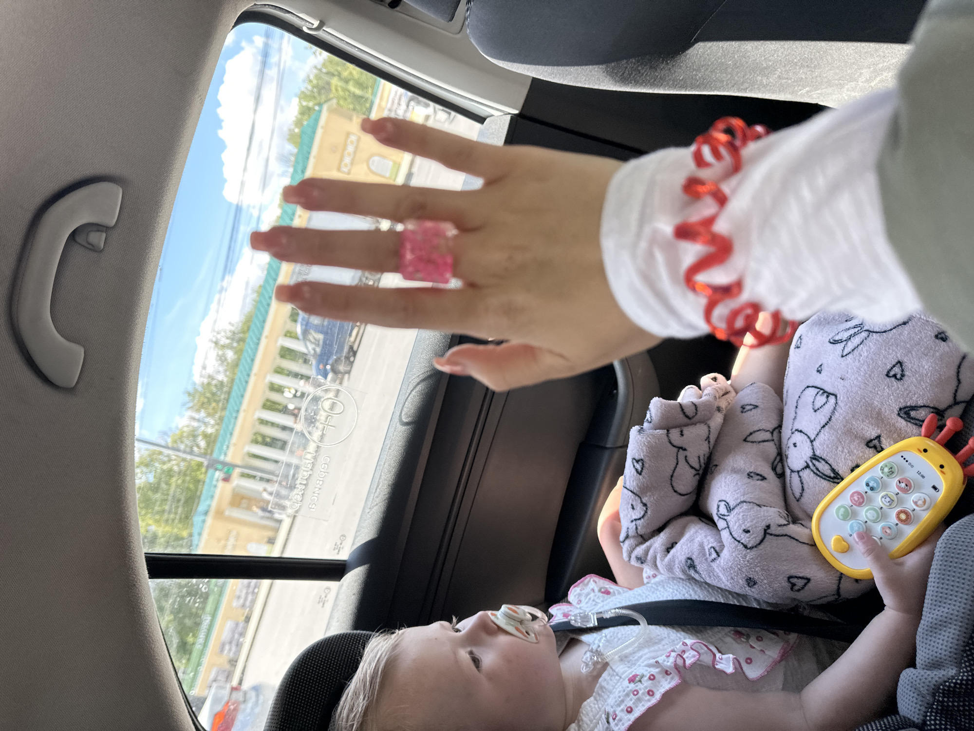 The image provides a view from inside a car on a bright, sunny day, looking out a side window. In the foreground, a baby or toddler is seated in a car seat, facing upward and to the left. The baby has light hair, a pacifier in its mouth, and is wearing a light-colored outfit with pink frills and small floral designs. A soft, pink blanket patterned with rabbits and hearts covers the baby's lap. The baby's right hand holds a yellow toy phone with colorful buttons and two red antennae.
In the upper right portion of the frame, an adult's hand is visible, extended forward. This hand wears a square, transparent pink ring with embedded material on the ring finger and a red spiral-shaped accessory (likely a hair tie or bracelet) on the wrist.
Outside the car window, a bright outdoor scene unfolds under a blue sky with scattered white clouds. A multi-story building with a light yellow or tan facade, a green roof, and white architectural elements such as columns or balustrades is visible. Trees and a partially visible dark blue car are also seen outside.
The car's interior includes a gray ceiling lining and a grab handle. The time of day is clearly daylight, indicated by the bright sun and clear sky. Visible text on the car window glass includes "сведения" (meaning "information" or "details") and "0+", indicating an age restriction often found on child safety equipment, consistent with the provided location context of Nizhny Novgorod, Russia. The baby appears calm and engaged with its toy.