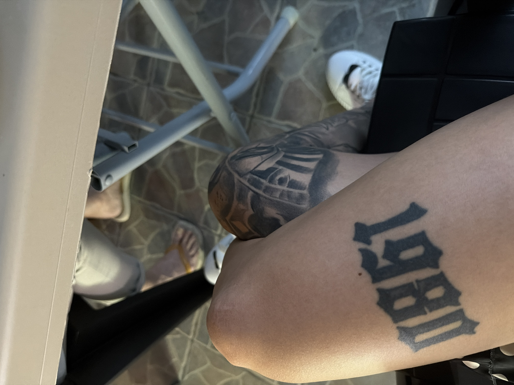 The image shows a close-up view of a person's leg with a tattoo of the number "0861" in a gothic font. The tattoo is dark and appears to be a recent addition. In the background, there are glimpses of other people's legs and feet, one wearing yellow flip-flops and another wearing white sneakers. There is also a metal folding table and a tiled floor visible. The scene appears to be outdoors, possibly in a cafe or restaurant setting, though there is no specific indication of the city of Plovdiv, Bulgaria, within the image itself. The lighting is natural, suggesting daytime.