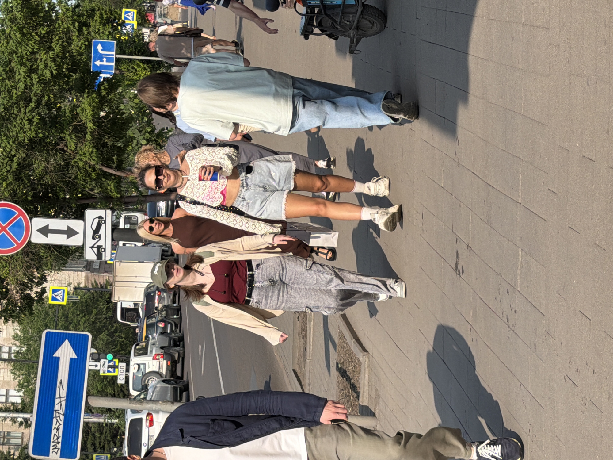 On a sunny day, several people are walking on a sidewalk. A woman in the foreground wears a light-colored cropped top and denim shorts, holding a cup. Next to her, another woman wears a beige jacket over a burgundy top and patterned pants, with a green baseball cap. A man in a dark jacket and light pants walks on the left, with his phone in his hand. In the background, traffic is visible on a street, including cars and trucks. The setting appears to be an urban environment with trees and buildings lining the street. The image was captured on Komsomolsky Avenue in Krasnoyarsk, Russia, based on the presence of a street sign with "КОМСОМОЛЬСКИЙ ПРОСПЕКТ" inscribed on it.