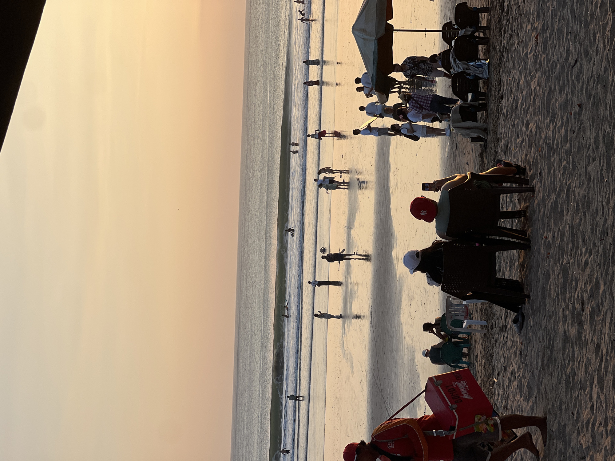 This is a photograph of a beach scene at sunset in Kuta, Indonesia. The sky is a warm gradient of orange and yellow, with the sun low on the horizon. The ocean is calm with gentle waves, and several people are visible in the water, some of whom appear to be surfing. The wet sand of the beach is reflective, showing the silhouettes of people scattered across it. In the foreground, a person wearing a red vest and carrying a red cooler walks across the sand. To the right, two people are seated in chairs under an umbrella, and other beachgoers are mingling or seated. The overall atmosphere is one of a relaxing evening on a popular tourist beach.