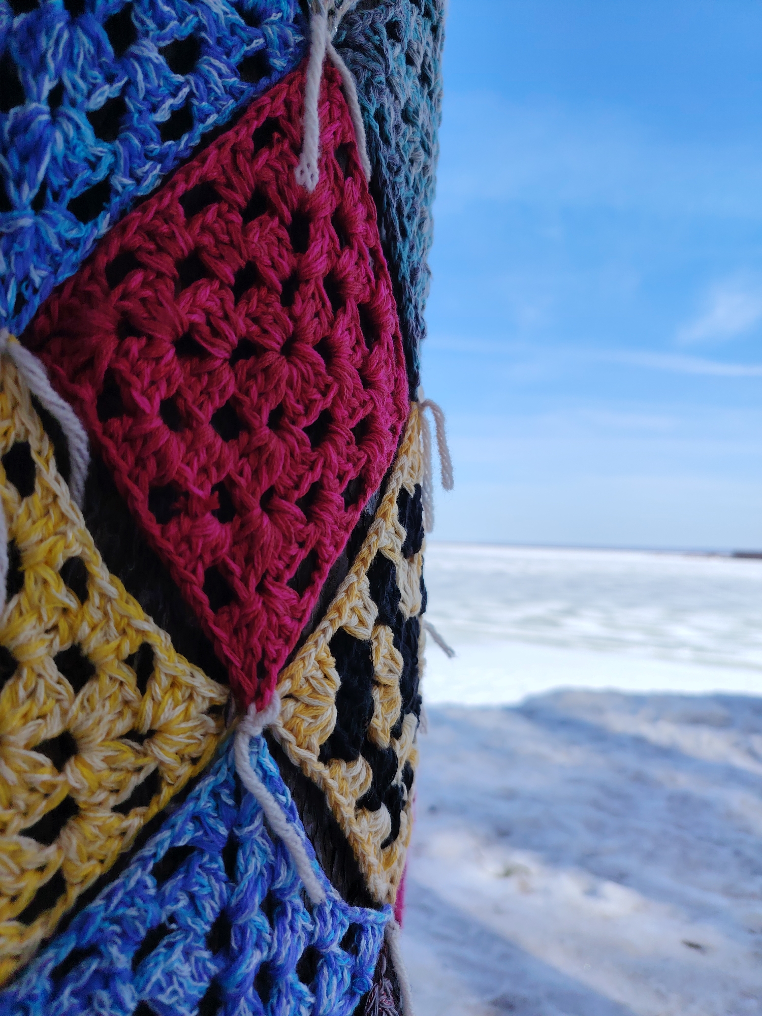 The image shows a close-up view of a tree trunk adorned with colorful, crocheted granny squares. The squares are various colors, including blue, yellow, pink, and black. White yarn ties secure the squares to the tree. In the background, a snow-covered landscape stretches out to a partially frozen body of water. The sky is blue with wispy clouds. The photograph was taken in the city of Chaikovsky, Russia.