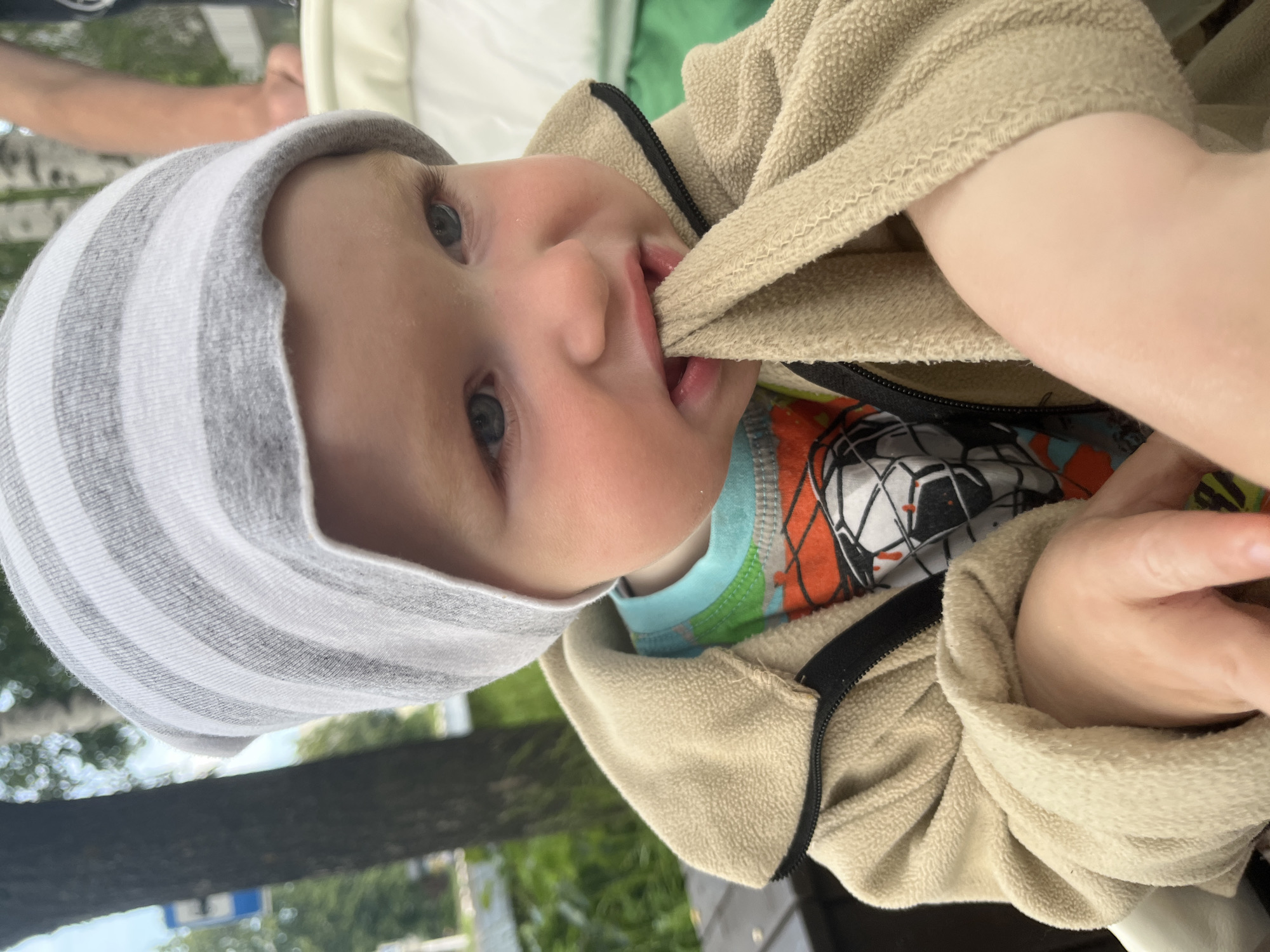 A close-up, eye-level shot shows a young Caucasian child in Chaikovsky, Russia, wearing a gray and white striped beanie and a tan fleece jacket zipped up. The child is chewing on the fabric of their jacket sleeve, with their mouth partially open. They have light skin, light eyebrows, and blue eyes looking to the right. Underneath the jacket, a t-shirt with a colorful graphic is visible. The background is blurred, with hints of trees, foliage, a possible sign, and the back of a person's arm. The lighting suggests it is daytime, likely outdoors.