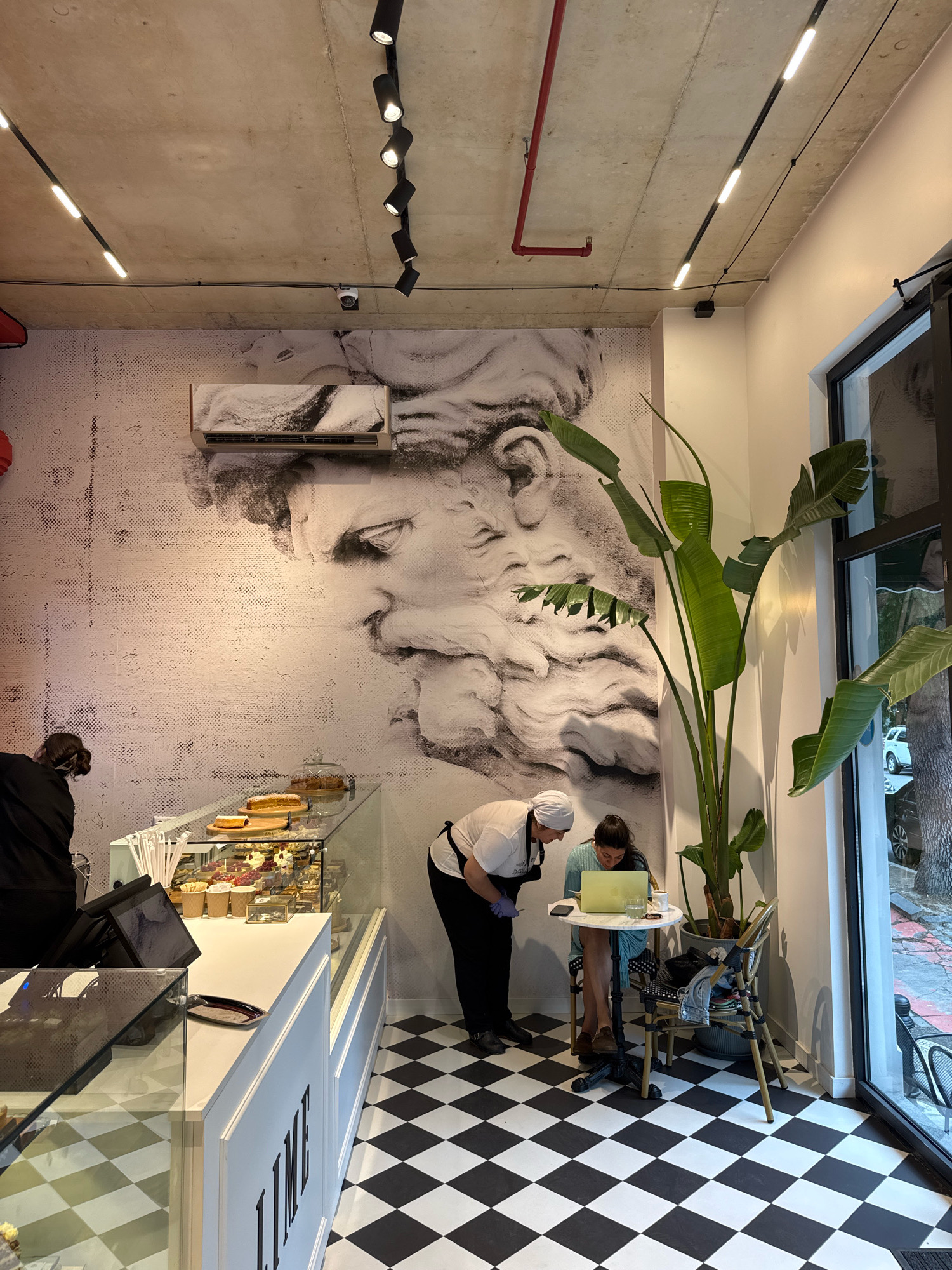 This is an indoor scene of a cafe with a black and white checkered floor. The wall features a large black and white mural of a man's profile. In the foreground, a display case filled with pastries is visible. To the right of the display case, two women are seated at a small table. One woman, wearing a light blue shirt, is looking at a laptop. The other woman, wearing a white headscarf and apron, appears to be speaking to her. A large potted plant stands beside the table. The cafe is well-lit with overhead track lighting. This cafe is located in Batumi, Georgia, as indicated by the text "LIME" visible on the display case, which is the name of the cafe.