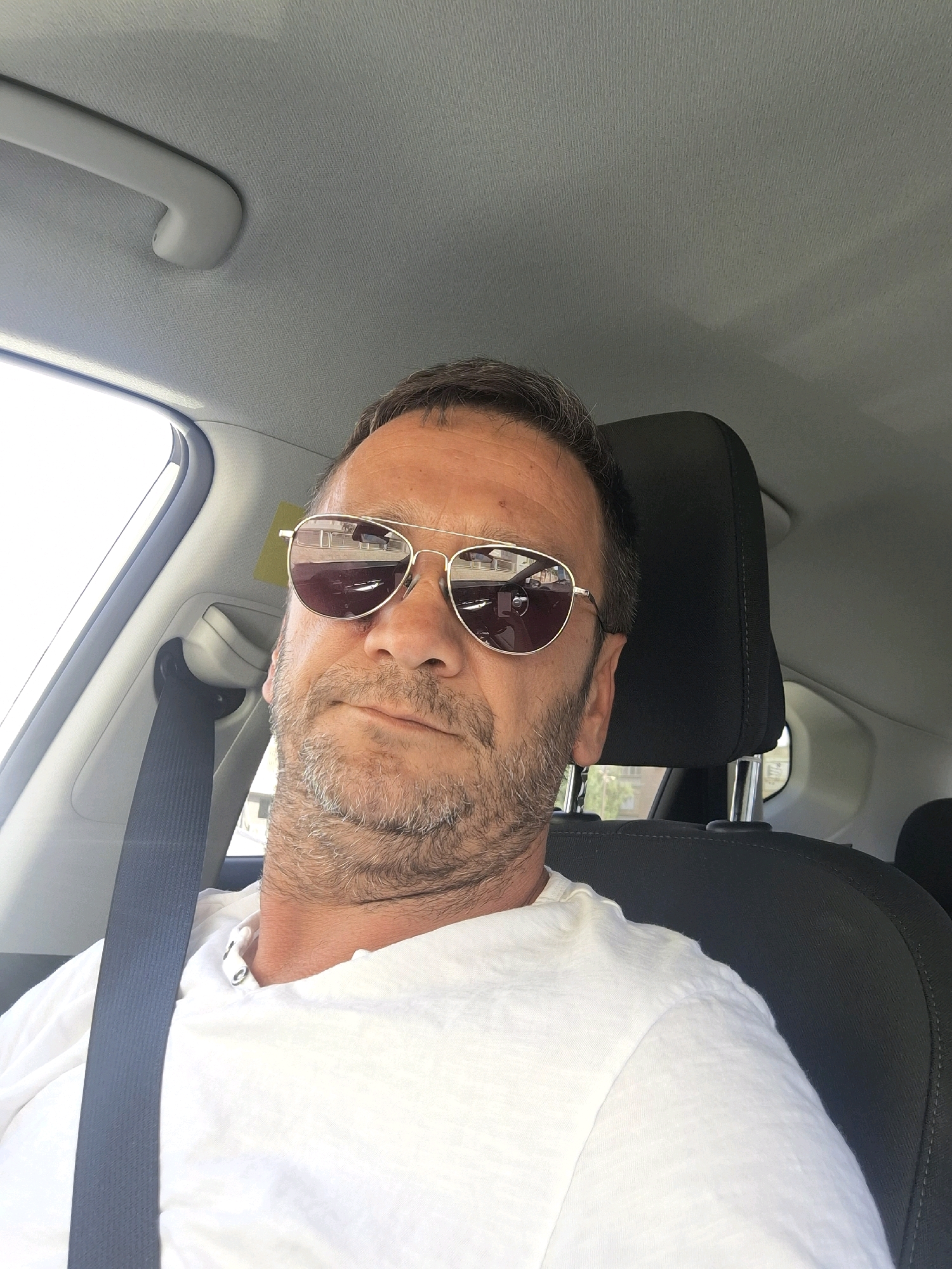 A man wearing sunglasses and a white t-shirt is sitting in the passenger seat of a car. The car appears to be parked, as indicated by the light reflecting on the sunglasses, suggesting the man is looking out the window. The interior of the car is visible, including a black seat and a gray headliner. A seatbelt is fastened across the man's chest. The lighting suggests it is daytime. The image was taken in Dimona, Israel.