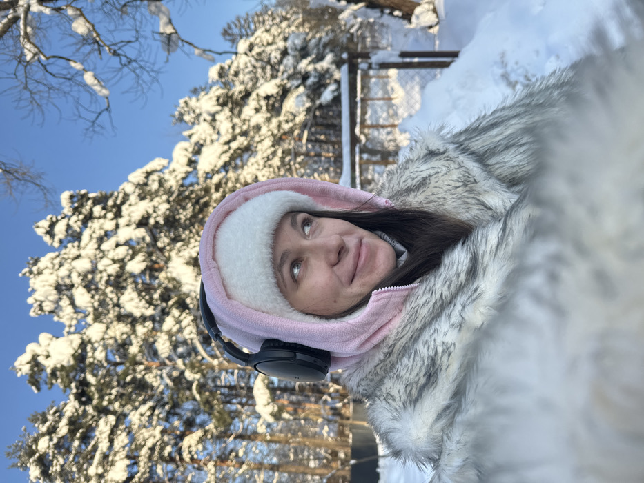 A young adult female is depicted outdoors in a snowy environment, identified as Дубки, Russia. She is shown from the upper chest upward, looking towards the upper right of the frame with a slight smile. She wears a white, furry winter hat under a pink hooded fleece jacket, with black over-ear headphones placed on top of the hood. A grey and white faux fur coat is visible on her shoulders. Her dark hair extends beneath the hat. The background features a deeply snow-covered landscape under a clear blue sky. Numerous coniferous trees, heavily laden with snow, dominate the mid-ground and background. Bare deciduous tree branches, also dusted with snow, are visible on the right. A wooden fence, partially obscured by snowdrifts, is present in the middle ground to the right. The ground is uniformly covered in deep, white snow, with shadows suggesting bright sunlight.