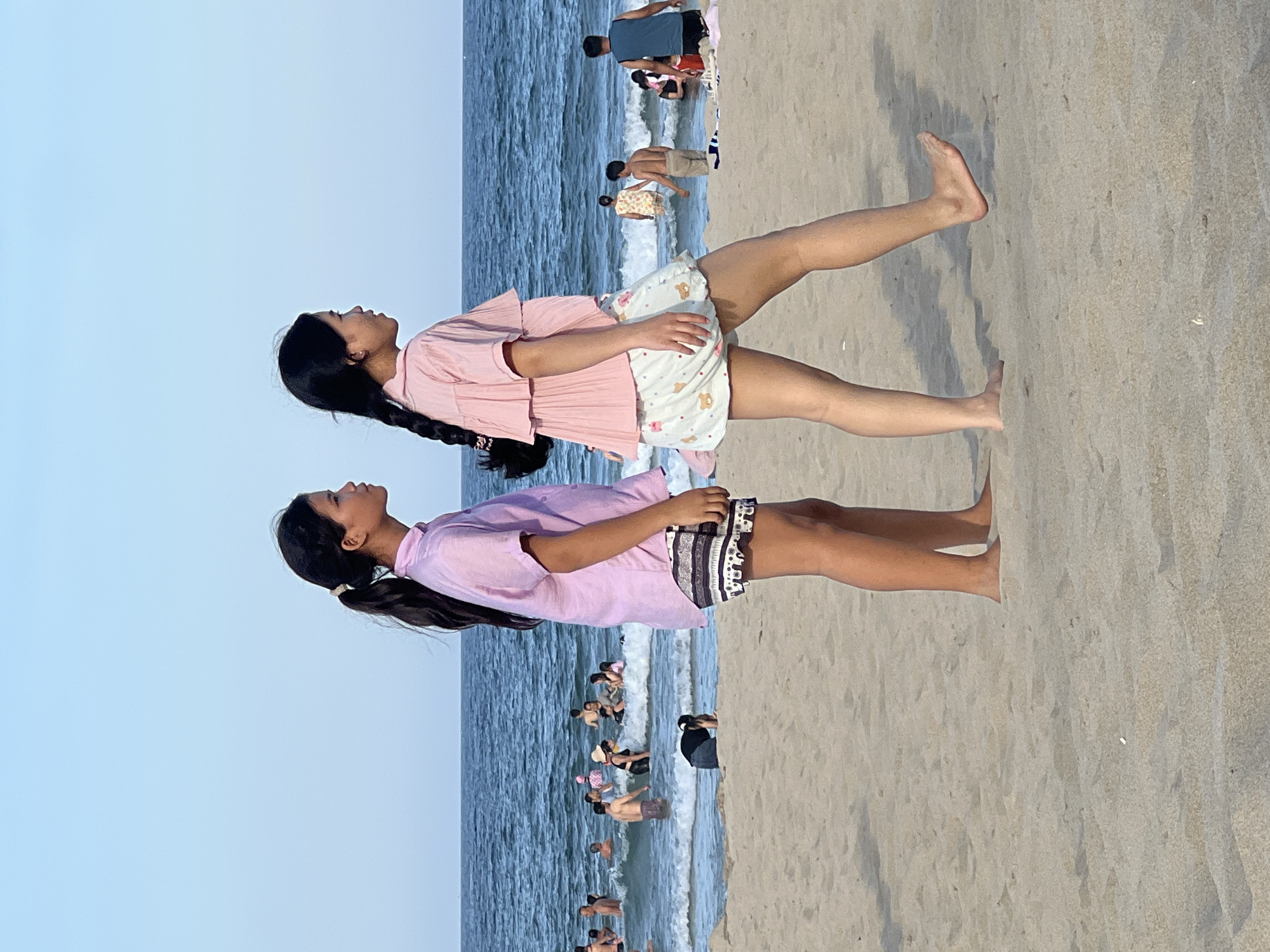Two young females, likely of Asian descent, are walking on a sandy beach. The female on the left wears a lavender short-sleeved shirt and patterned shorts. Her dark hair is pulled back into a ponytail. The female on the right wears a light pink short-sleeved shirt with ruffled sleeves and white shorts with a pattern of small bears. Her dark hair is in a braid. Both are walking from left to right in the frame, facing towards the right.
In the background, the ocean is visible with moderate waves. Numerous people are in the water, swimming and wading. Some are standing, while others are partially submerged. The sky is clear and blue. The beach is composed of light-colored sand.
The scene appears to be a public beach during daylight hours, likely a popular tourist destination in the city of Đà Nẵng, Vietnam, given the presence of diverse individuals enjoying the ocean.