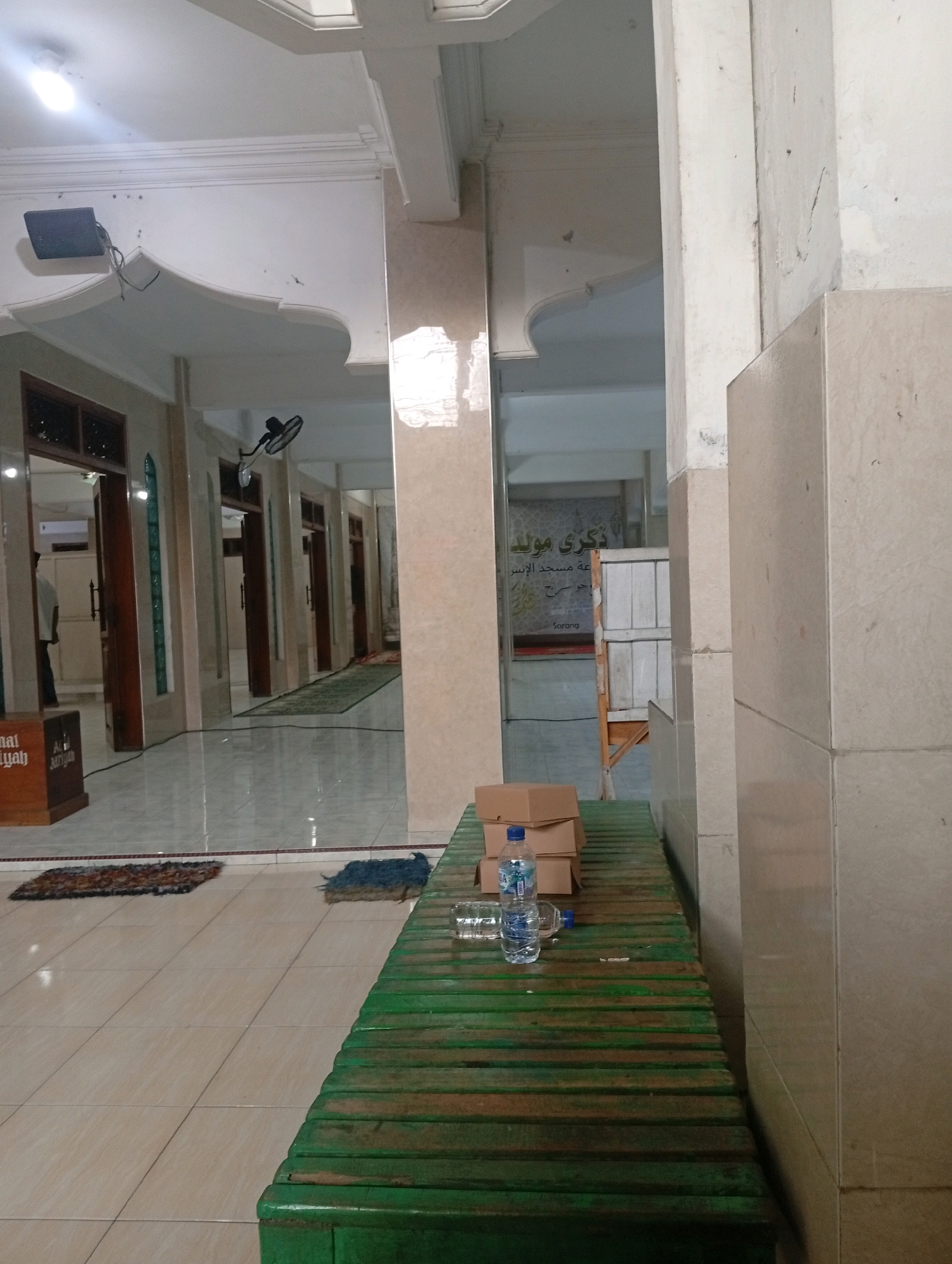 This is a photograph taken indoors, likely within a mosque in Bajingjowo, Indonesia, given the presence of Islamic calligraphy on signage and the general architectural style. The scene depicts a long, narrow hallway with tiled flooring and beige-colored tiled walls. On the left side of the hallway, there are several open doorways leading to other rooms. A person, partially visible, stands in one of these doorways. Along the hallway, there are green prayer mats laid out on the floor, with one blue mat near the foreground. In the foreground, a weathered green wooden bench is positioned diagonally across the frame. On the bench, there are several stacked cardboard boxes and a bottle of water. The ceiling is white, with recessed lighting fixtures visible. The overall impression is one of a functional and somewhat aged interior space.