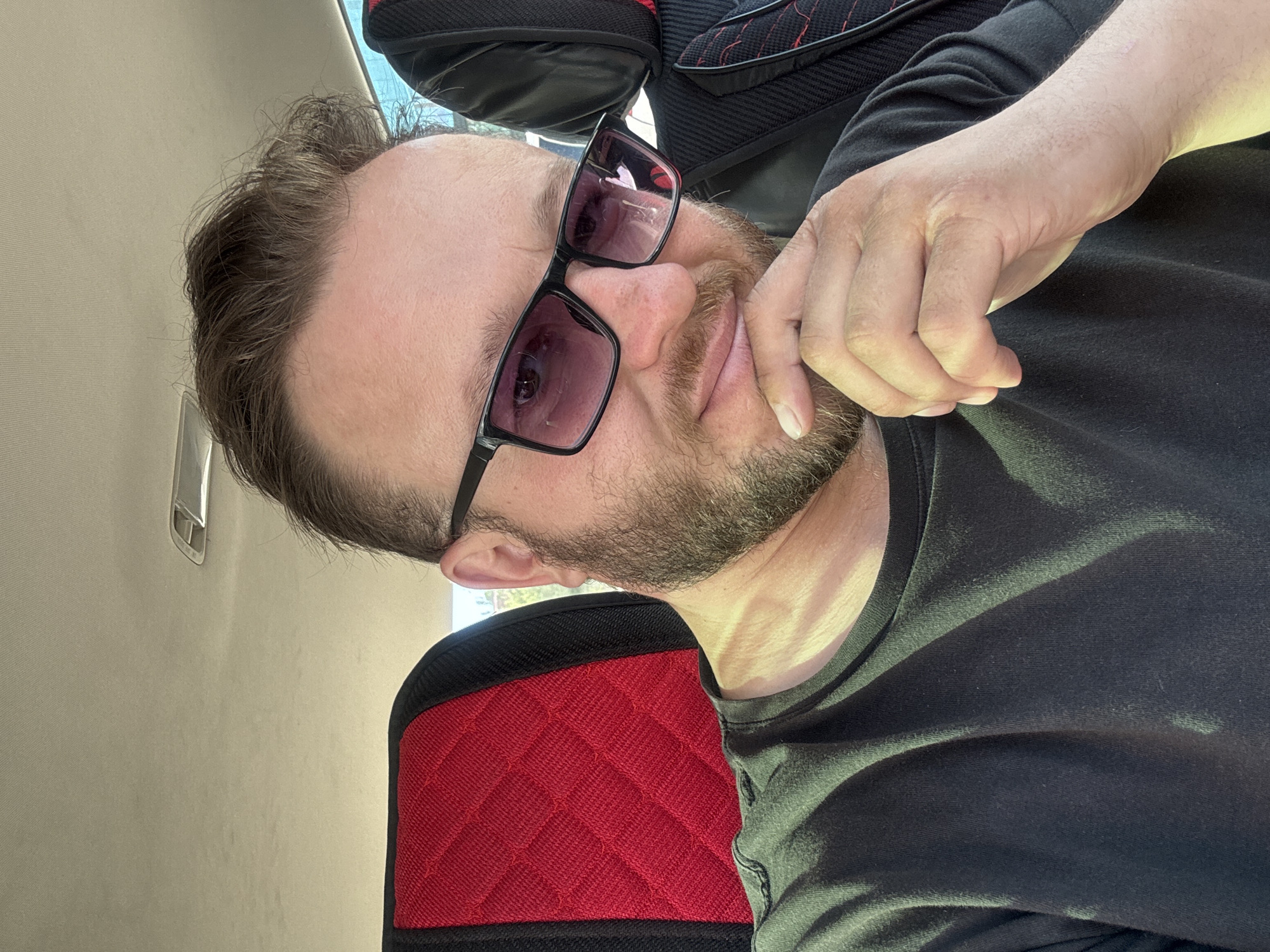 A man wearing sunglasses and a black t-shirt is seated in a car. The car interior features red and black patterned upholstery on the headrests. The man has short, brown hair and a beard. He is resting his chin on his hand and looking at the camera. The overall lighting suggests it is daytime. The image was taken in Blagoveshchensk, Russia.
