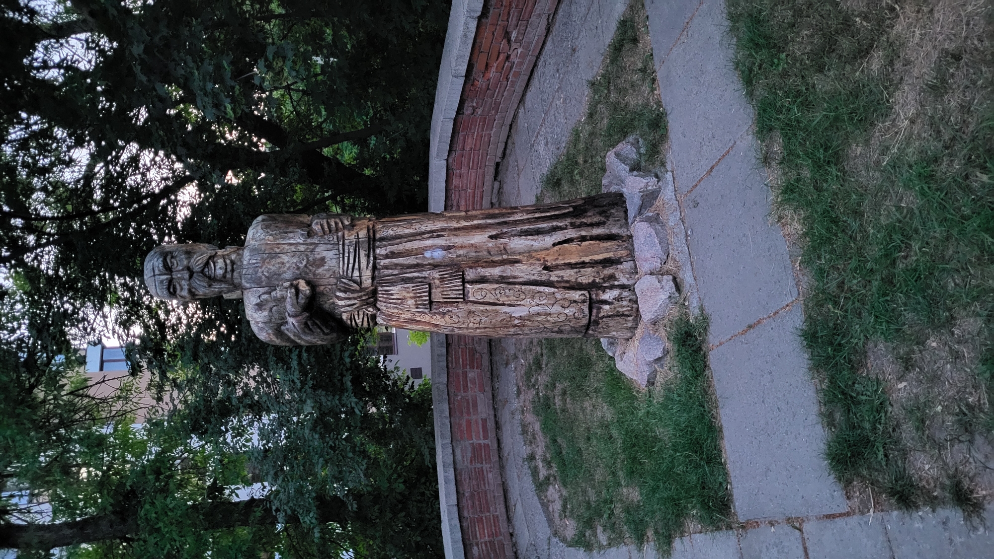 The image depicts a vertical wooden sculpture of a person, possibly a man, situated outdoors. The sculpture is carved from a rough-hewn log, showing visible grain and texture. The figure appears to be wearing a robe or tunic, with carved details on the chest and sleeves. The head is also carved with facial features, including a beard and eyes, giving it a wise or elderly appearance. The sculpture is positioned on a paved area, with grass and some rocks surrounding its base. In the background, there are trees with green foliage, suggesting a park or garden setting. A brick wall curves around part of the paved area, and a building with windows is partially visible through the trees. The lighting suggests it is either early morning or late afternoon, with the sun filtering through the trees, creating a somewhat subdued atmosphere. The overall scene is serene and contemplative, focusing on the handcrafted sculpture and its natural surroundings.