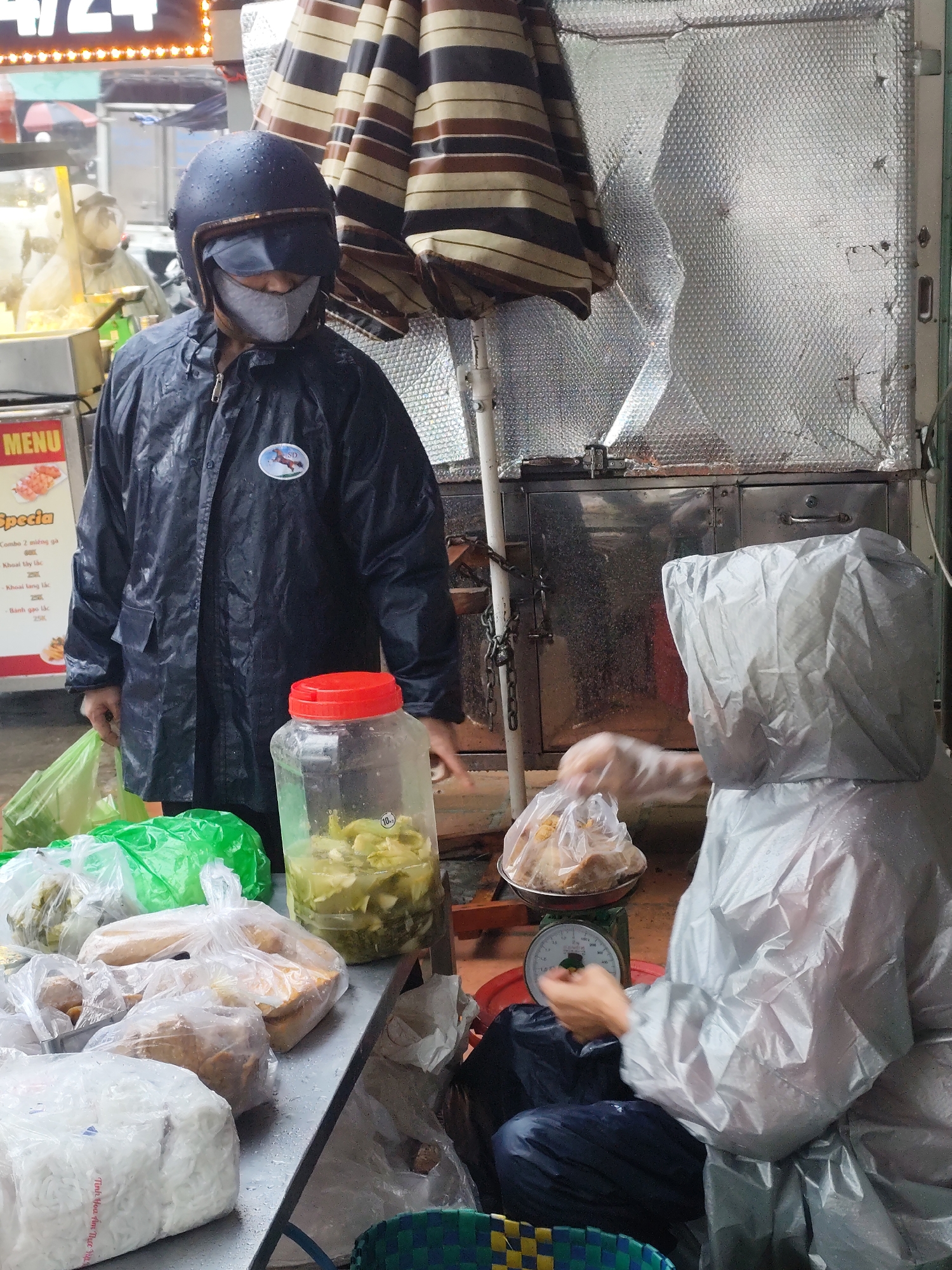 In a street scene, likely in Lang Biang - Đà Lạt, Vietnam, two individuals are involved in a transaction at a street food stall during a rainy day. The first individual, standing to the left, is wearing a dark raincoat and a helmet with a face mask. A sticker is visible on the raincoat. This person is looking towards the stall. In front of them is a table with several plastic bags containing food items, including what appears to be rice noodles and fried items. A large jar filled with pickled vegetables sits near the edge of the table.
The second individual is seated and covered almost entirely in a large, opaque rain poncho. They are weighing food items in a silver bowl on a green scale. The background includes a food preparation area with stainless steel surfaces and an umbrella providing some cover. A menu board with Vietnamese script and prices is visible, and there is a faint glow from an overhead sign with the numbers "4/24". The overall impression is of a busy but wet food vending scene.