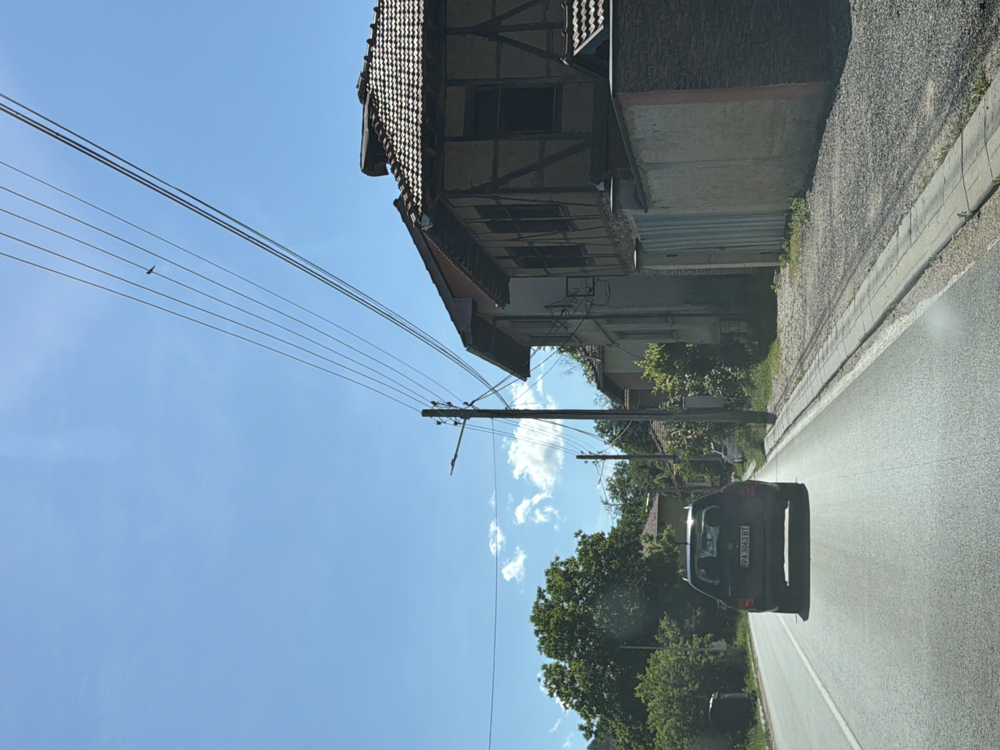 The image shows a road with a car driving away from the camera in the city of Beli Osam, Bulgaria. The sky is blue with a few white clouds. Overhead, power lines stretch across the scene, with a small bird perched on one of them. To the right of the road, there is a building with a tiled roof and a distinctive timber-framed facade. Further down the road, more houses and trees are visible. The car, a dark-colored hatchback, has a license plate that reads "PA 3063 BT". The road surface is asphalt, and there is a concrete curb and a gravel shoulder along the side. The overall scene depicts a rural or suburban street in daylight.