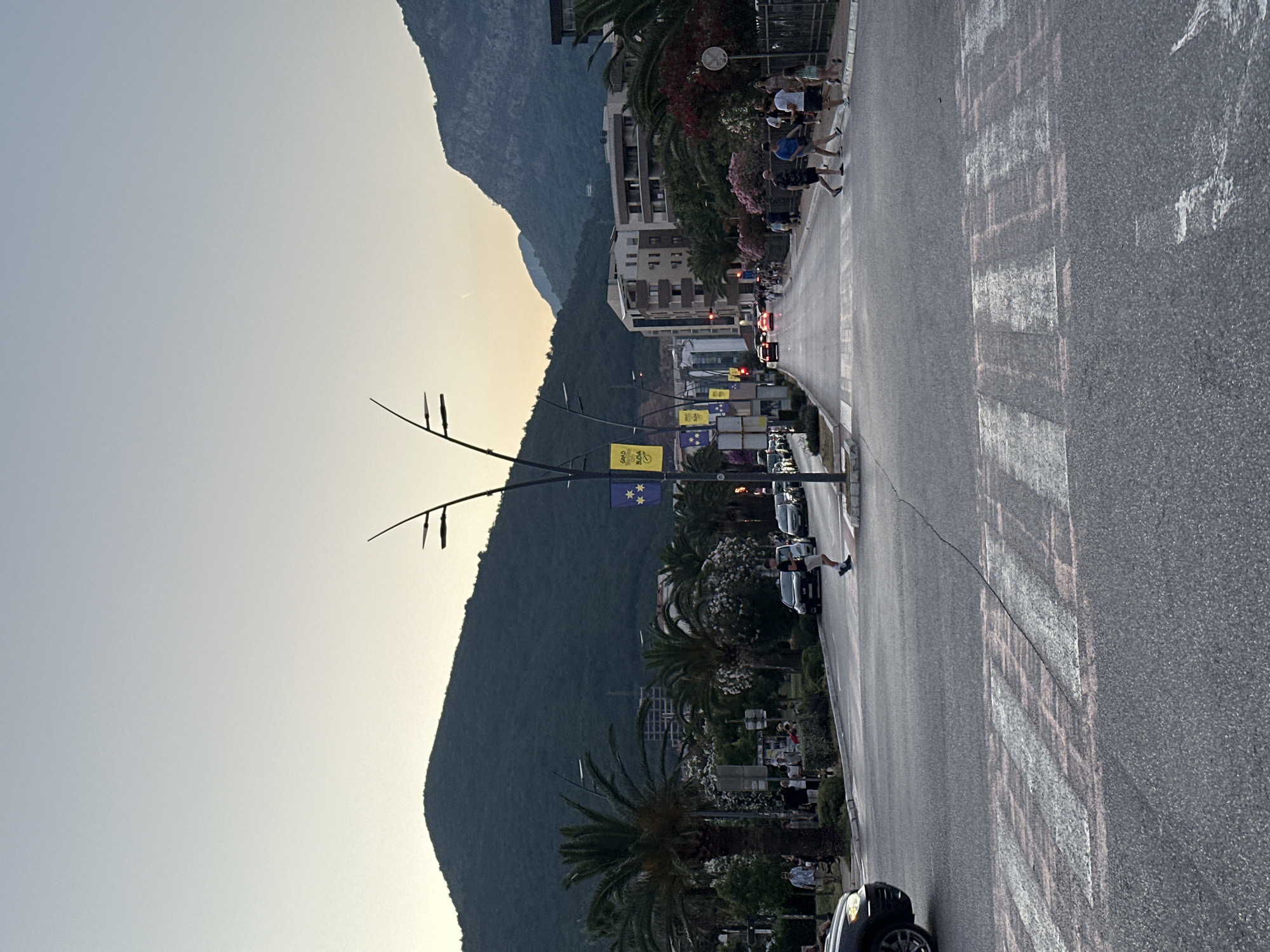The image depicts a street scene in Budva, Montenegro, likely during the late afternoon or early evening. A wide asphalt road stretches into the distance, flanked by sidewalks and buildings. In the foreground, a pedestrian crosswalk is visible, with faint pink markings overlaid on the white stripes. Several cars are parked or driving on the road, and a few people are walking across the crosswalk. Palm trees and lush vegetation line the sides of the street, and in the background, mountains rise against a pale sky. Flags with a yellow background and blue stars hang from lampposts, suggesting a local or national display. The overall atmosphere is one of a pleasant, urban environment with natural elements.