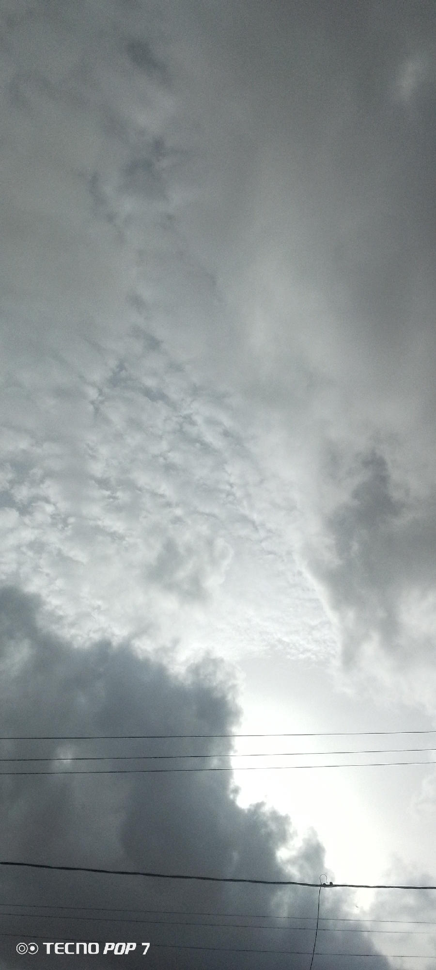 A low-angle shot captures a cloudy sky above power lines in Altagracia, Venezuela. Dark, heavy clouds dominate the lower portion of the frame, with patches of lighter, textured clouds above. A bright area of light, possibly the sun breaking through, is visible through a gap in the darker clouds. Four thin, dark power lines stretch horizontally across the lower third of the image, with one line dipping slightly and appearing to have a small object on it. The bottom left corner of the image displays the watermark "TECNO POP 7". The scene depicts an overcast or stormy sky, typical of many tropical regions.