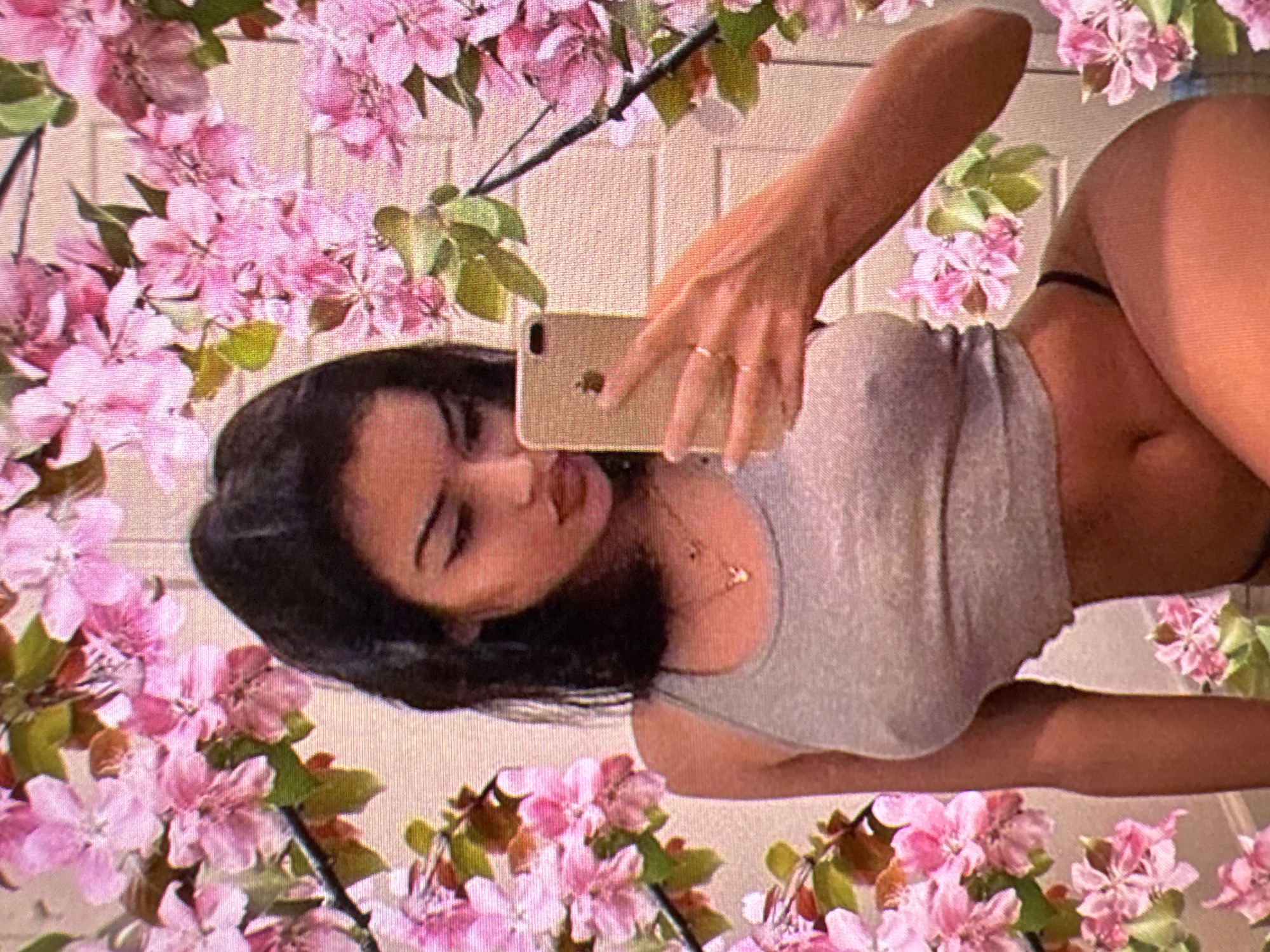 A young woman with dark hair is taking a selfie in front of a floral background. She is wearing a grey tank top and dark underwear. She is holding a gold iPhone in her right hand. The background appears to be a wall with a floral pattern, possibly wallpaper or a mural. The image is taken in Castelo Branco, Portugal.