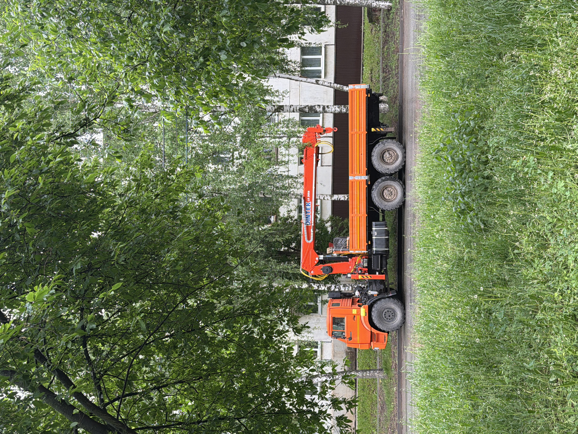 The image shows an orange truck with a crane attachment parked on the side of a road. The truck is positioned between a grassy shoulder and a tree-lined area, with a building visible in the background. The crane arm is extended upwards. The location appears to be a street in a residential area of Votkinsk, Russia, based on the architectural style of the building and the general environment.