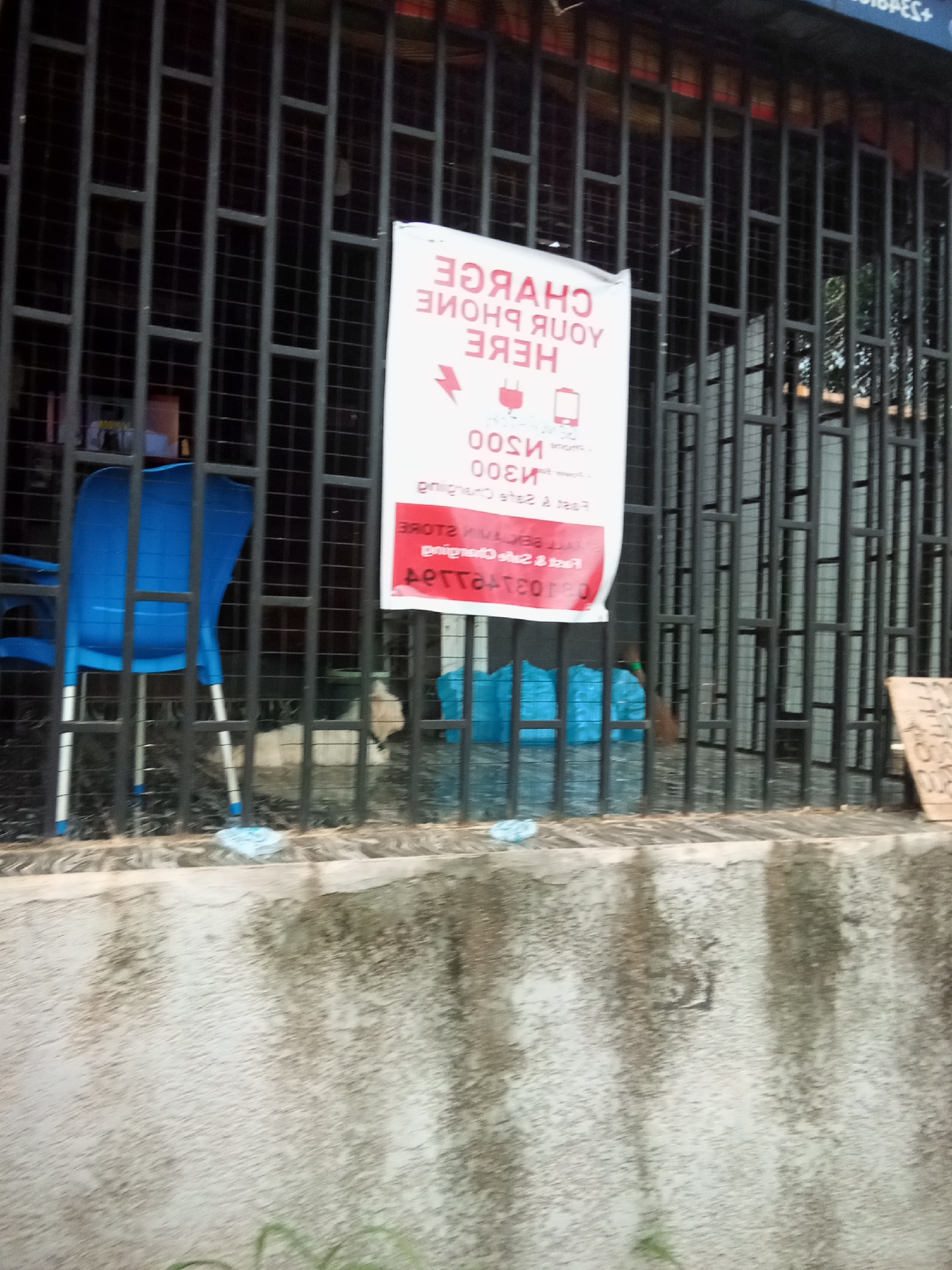 The image depicts a scene in Oko Town, Nigeria, characterized by a barred storefront. A white sign with red lettering, advertising "CHARGE YOUR PHONE HERE" is prominently displayed. Below this, the price of 300 Naira is visible. To the left of the sign, a blue plastic chair is positioned behind the bars. The ground in front of the storefront is paved with rough, textured concrete, showing signs of weathering and discoloration. In the background, a dark, barred gate suggests a secure or closed establishment. Further to the right, another sign, partly obscured, stands upright. The overall environment appears to be an outdoor commercial space, likely a street-side shop.