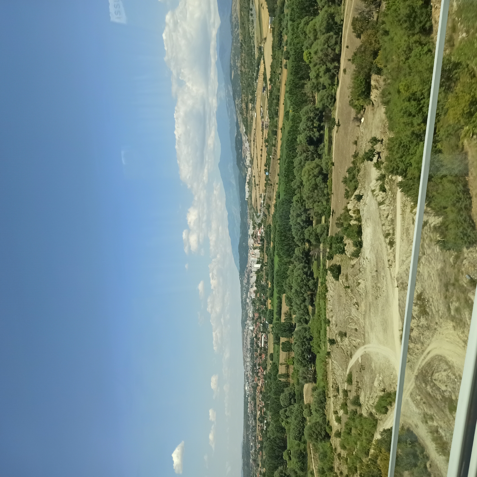 The image captures a wide-angle view of a landscape with a city in the distance, situated in Moshtanets, Bulgaria. The foreground features a dusty, arid terrain with winding dirt paths, bordered by a white railing. This area transitions into a lush green expanse of trees and vegetation, leading towards the more densely populated urban area. The city itself appears to be nestled in a valley, with buildings ranging from smaller residential structures to taller apartment complexes. Beyond the city, rolling hills and mountains form the horizon under a bright blue sky dotted with fluffy white clouds. The scene suggests a sunny day, possibly late morning or early afternoon. There are no people or animals visible in the frame.