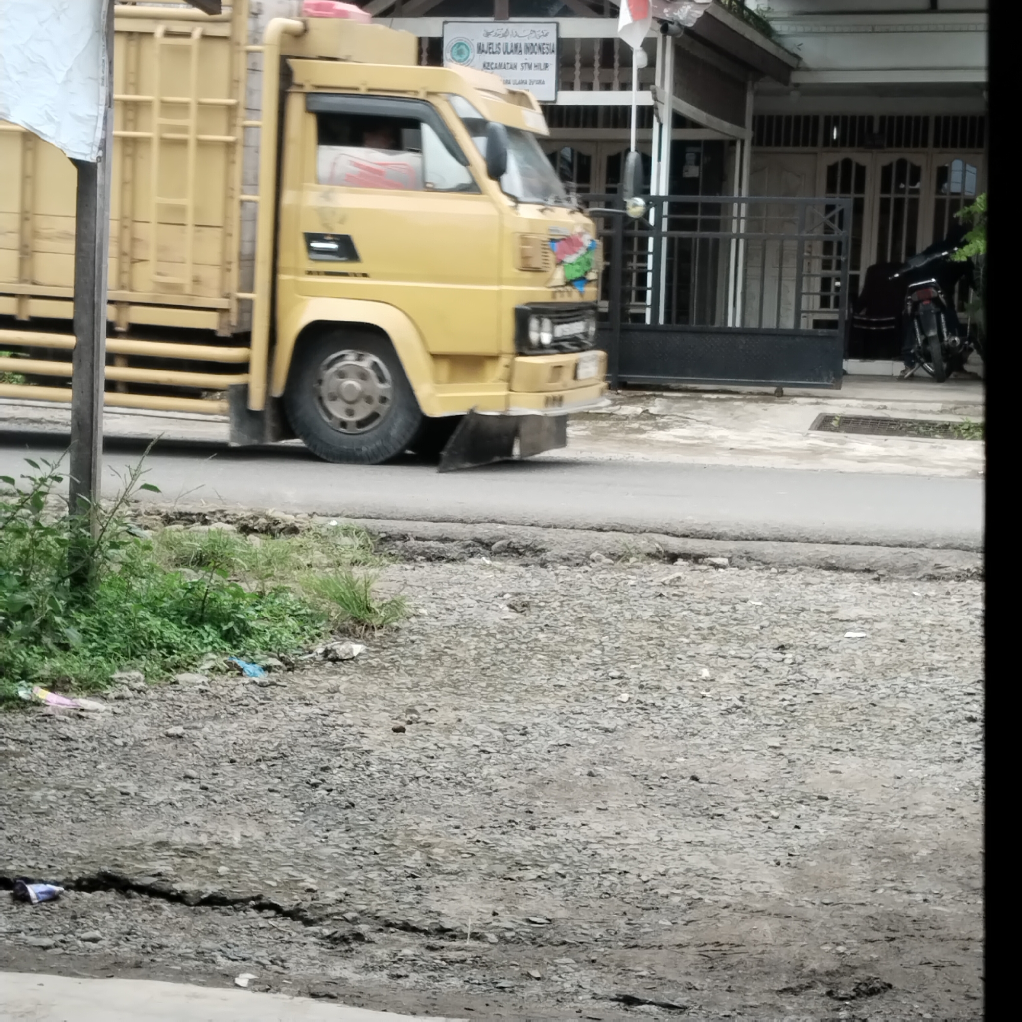 A yellow truck is driving past a building with a sign in Arabic and Indonesian that says "MAJELIS ULAMA INDONESIA KECAMATAN STM HILIR". A motorcycle is parked in front of the building. The ground is made of gravel and asphalt, with some overgrown weeds and scattered debris.