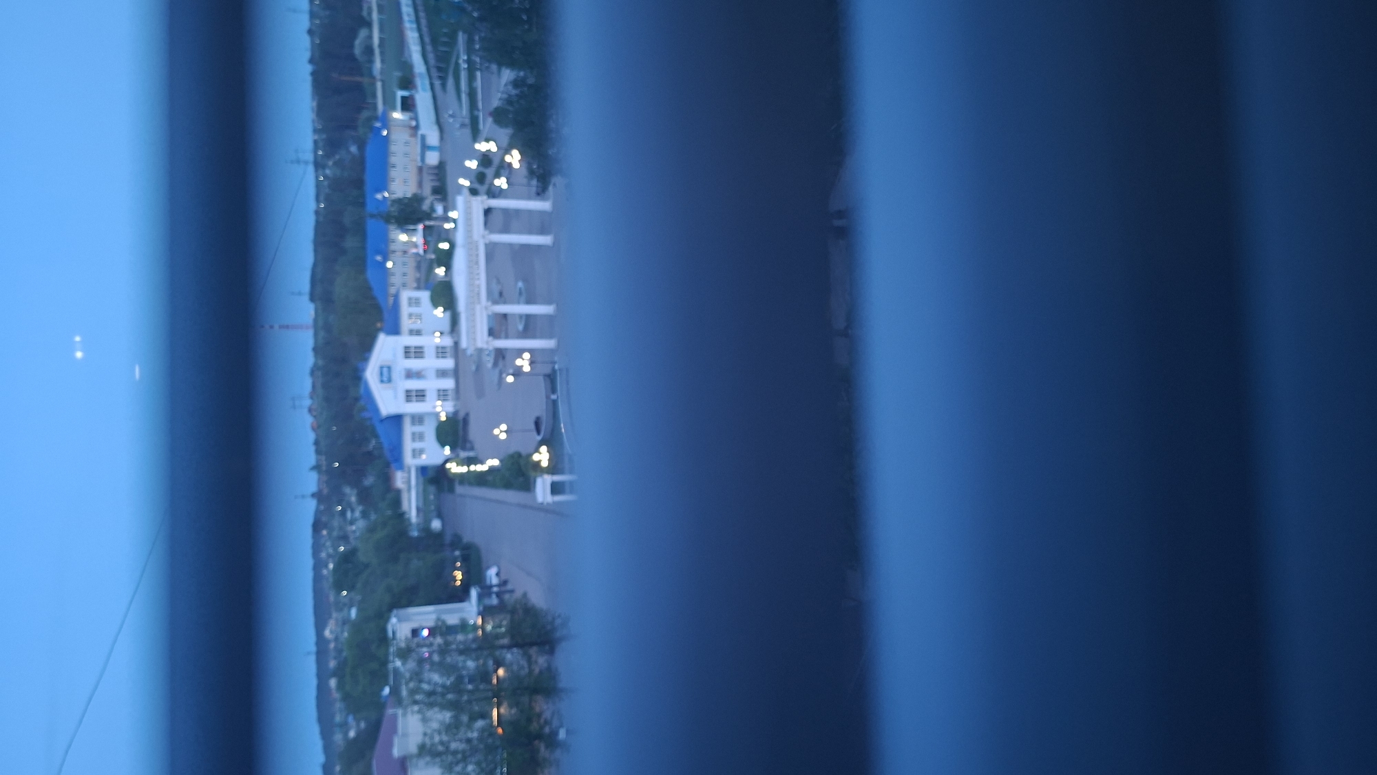 A low-angle, blurred outdoor photograph, possibly taken through a barred window, depicts a landscape in the city of Chaikovsky, Russia. The scene is dominated by a large, white building with a blue roof, located in the upper center of the frame. This building has multiple windows and appears to be several stories tall. A series of white columns extends from the building, creating an open arcade or walkway. Streetlights line a road leading towards the building. Further in the background, a body of water is visible, separating the foreground from a distant, dark strip of land. The sky is a clear, light blue. The focus is uneven, with the foreground and portions of the building rendered with low detail and blurring.