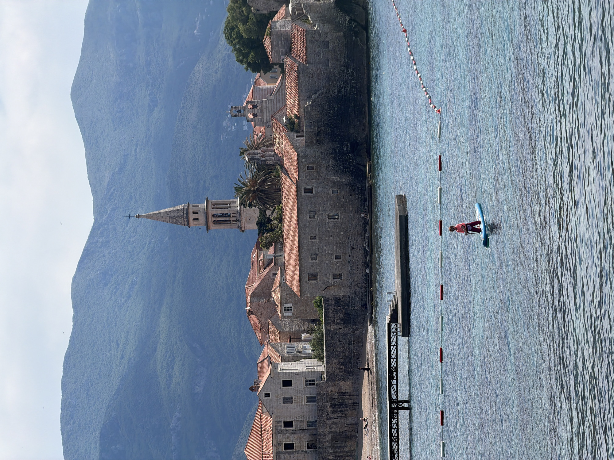 The image captures a scene in Budva, Montenegro, featuring the island of Sveti Stefan. Sveti Stefan is a fortified island village with terracotta-roofed buildings, churches, and a tall bell tower. Behind the island rises a mountain covered in dense vegetation. A woman is stand-up paddleboarding in the water, which is demarcated by a safety rope with red and white markers. A pier extends into the water near the paddleboarder. The sky is clear, suggesting a sunny day.