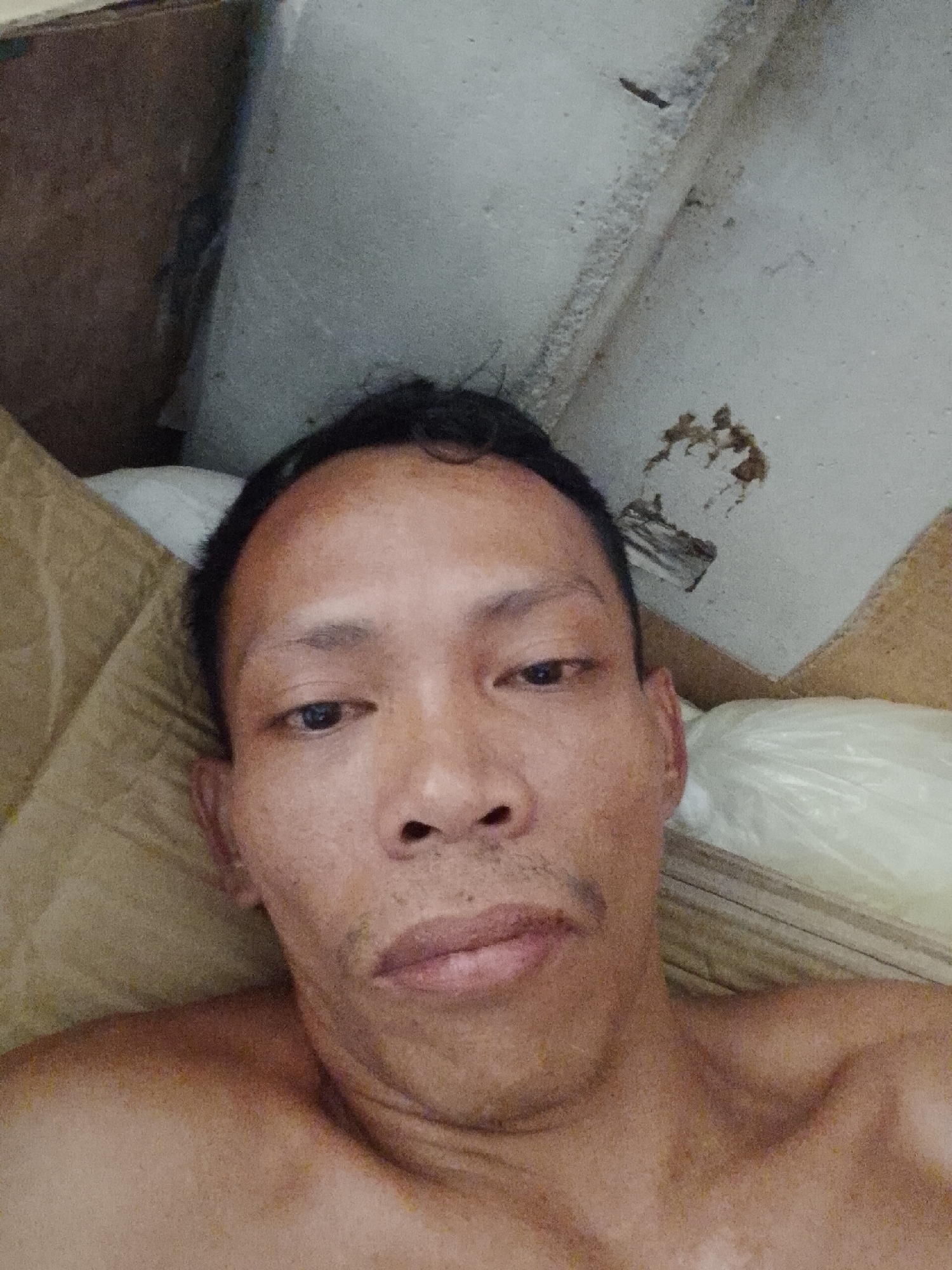 A man is lying down, appearing to take a selfie. He has short, dark hair, brown eyes, and dark skin. He is shirtless. He is surrounded by what appears to be cardboard and a white plastic bag. The location is Bukit Ketok, Indonesia. The walls appear to be made of concrete or cement with some discolored patches.